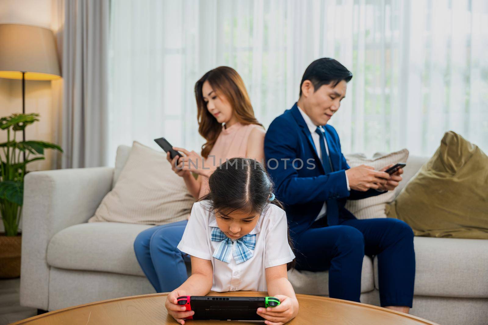 Family don't care about each other. Asian parents ignore their child and looking at their mobile phone at home, father and mother read social media but daughter play video game on sofa living room
