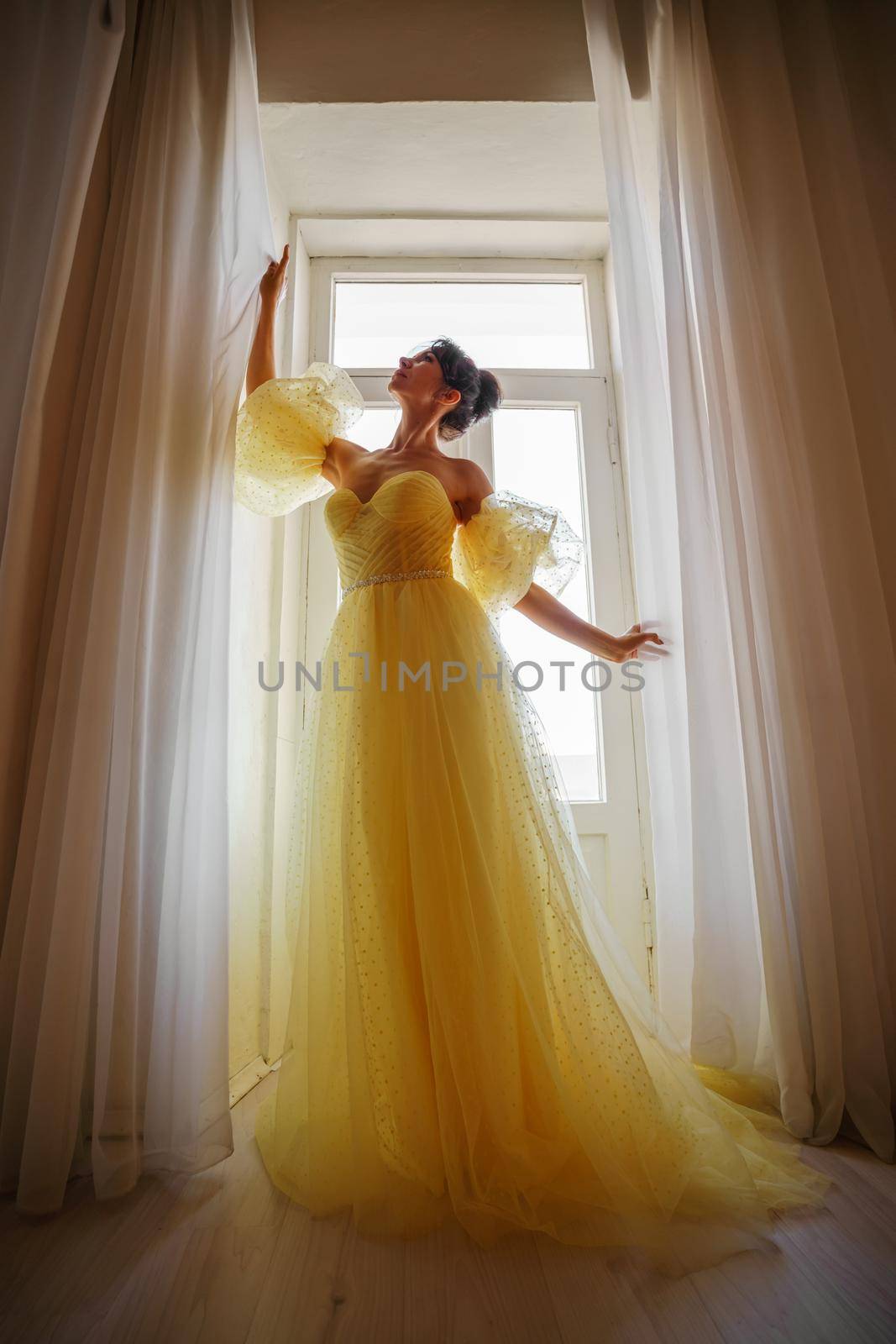 A woman's silhouette in a golden luxurious dress against the background of a window holds a curtain with her hands. Elegant lady in a yellow long silk dress with bare back, back view. by Matiunina