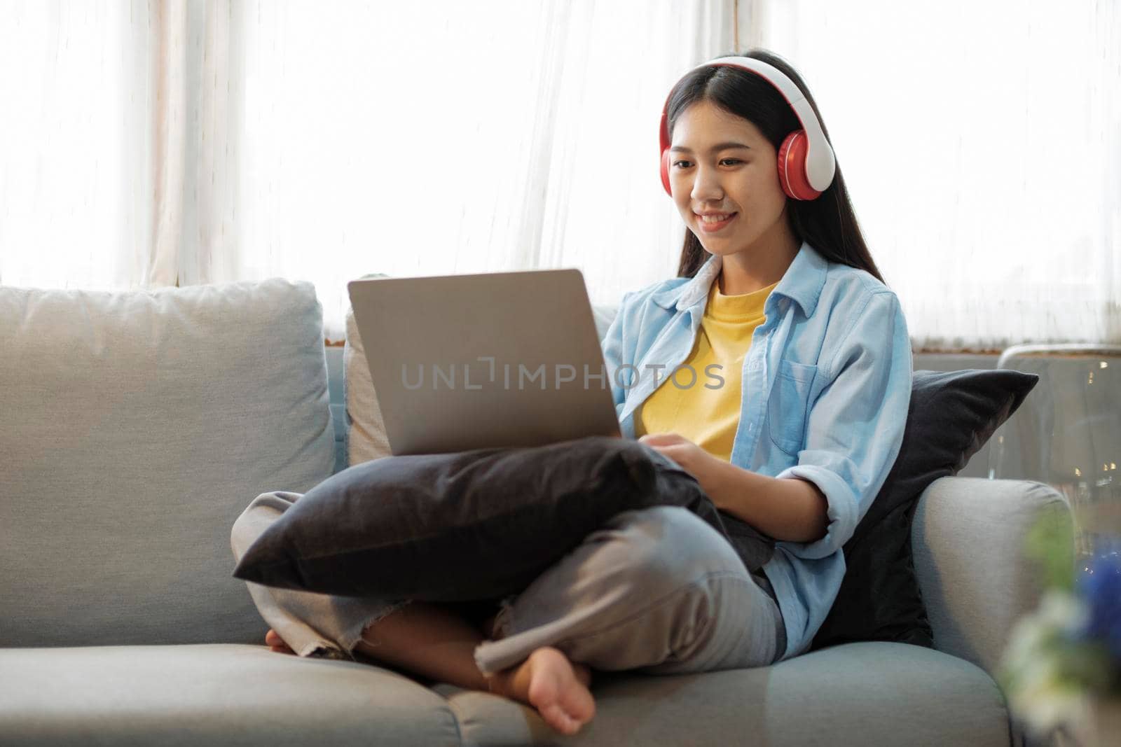 Young asian woman smiling listening to music using laptop surfing the internet while sitting on couch at home. Online connection and online learning concept.