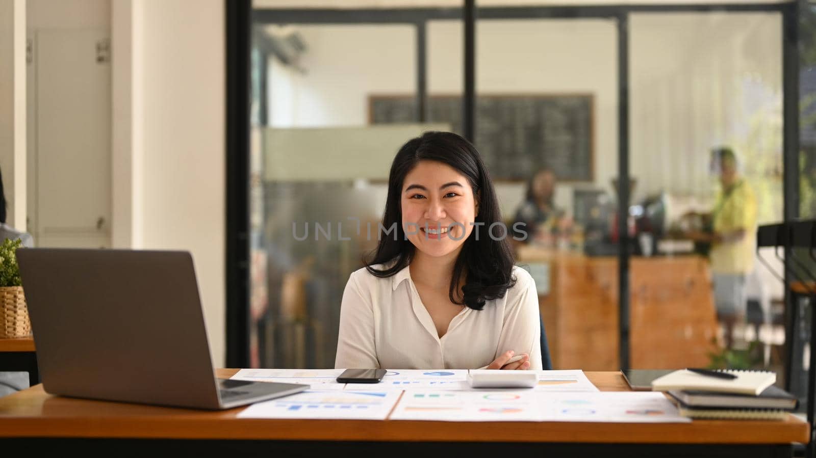 Cheerful asian female employee sitting with arms crossed at her office desk and smiling at camera.