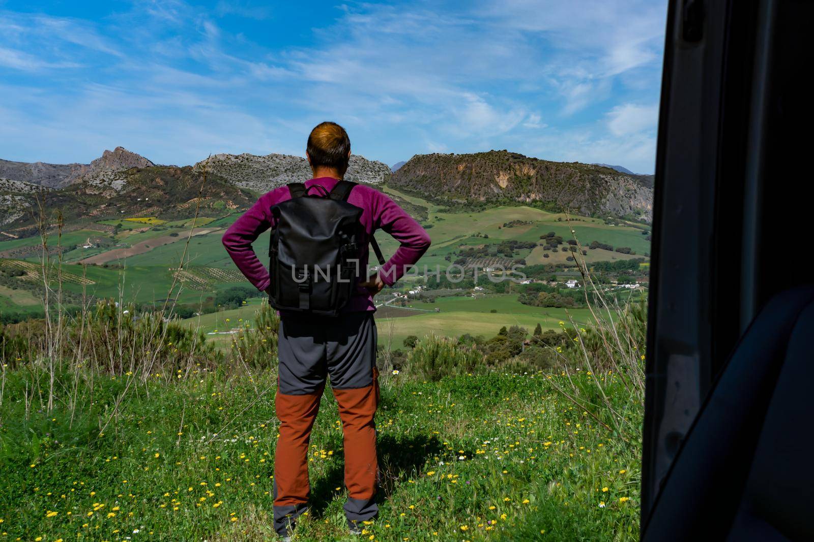 man on his back with backpack observing the mountainous landscape with cloudy sky seen from inside his van