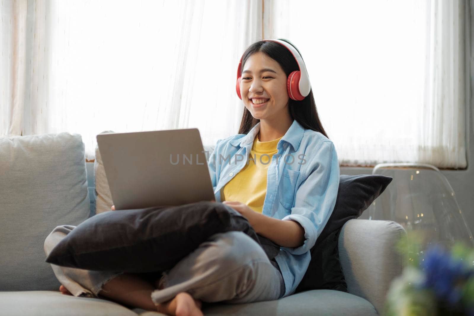 Young asian woman smiling listening to music using laptop surfing the internet while sitting on couch at home. Online connection and online learning concept.