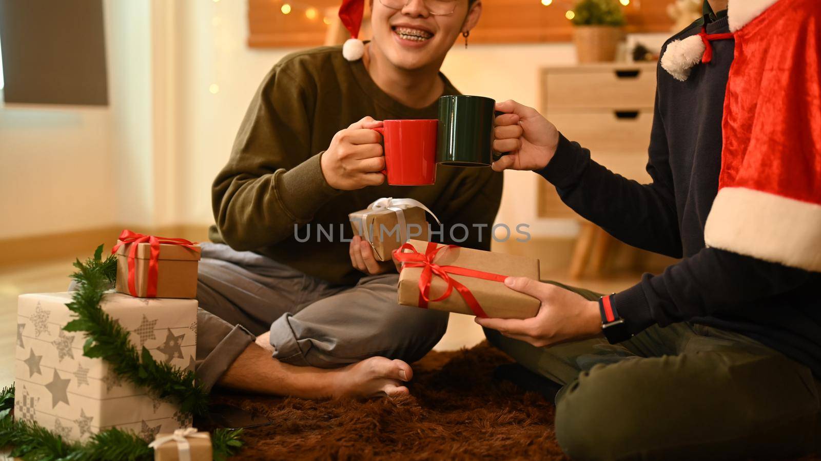 Two happy men clinking cups of hot chocolate while celebrating Christmas or New Year together in cozy living room.