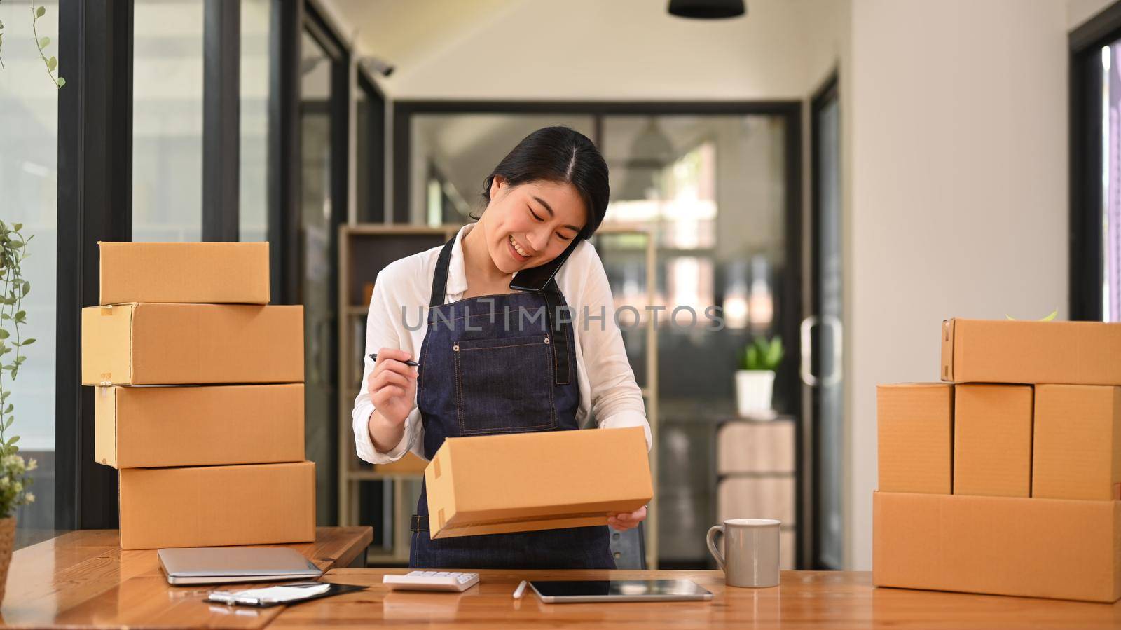 Female online seller in apron talking on mobile phone for confirming orders or taking orders from customer.