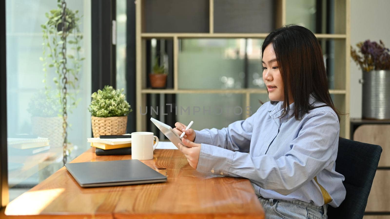 Asian woman office worker in blue shirt sitting at her workplace near window and using digital tablet.