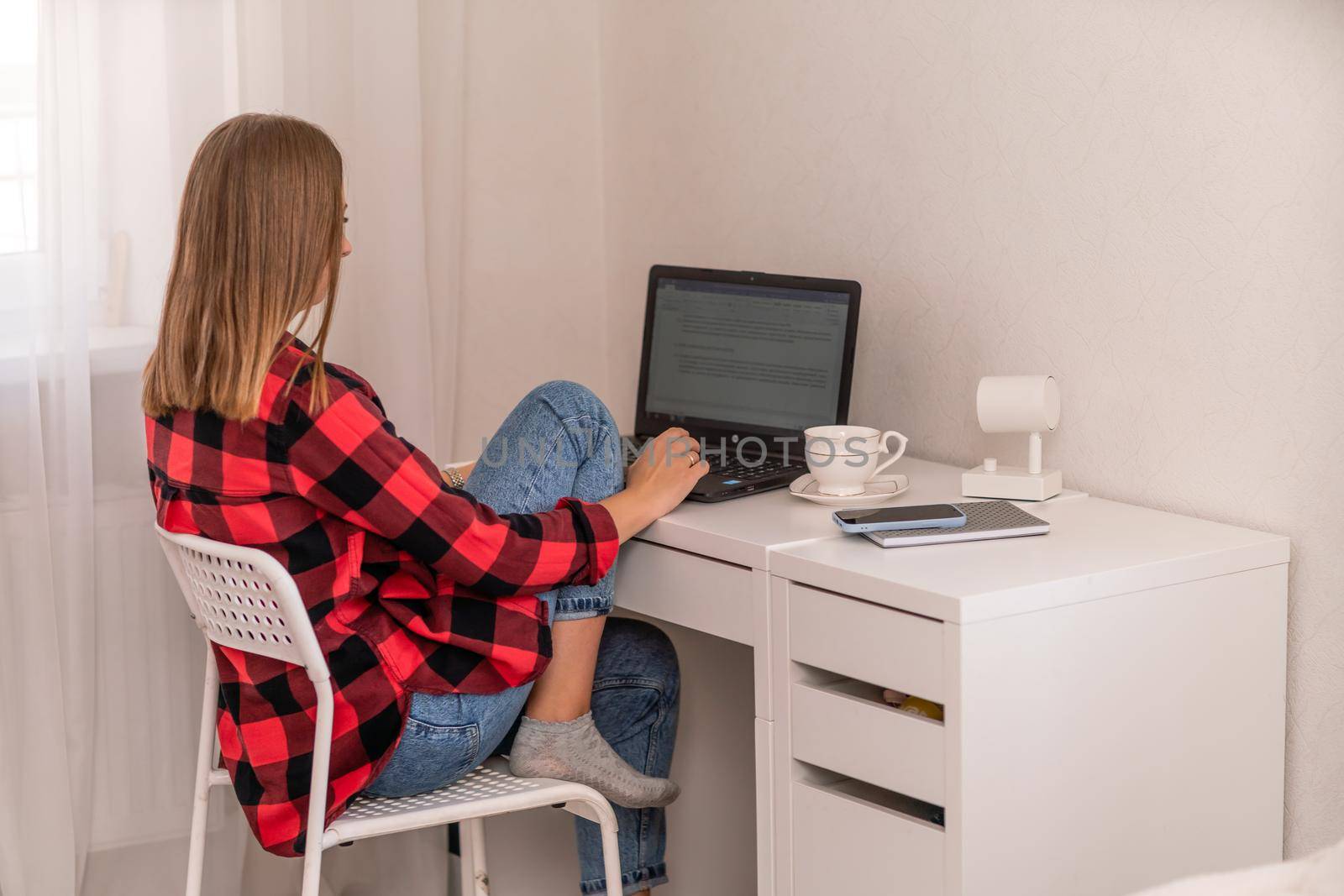 European professional woman sitting with laptop at home office desk, positive woman studying while working on PC. She is wearing a red plaid shirt and jeans. by Matiunina