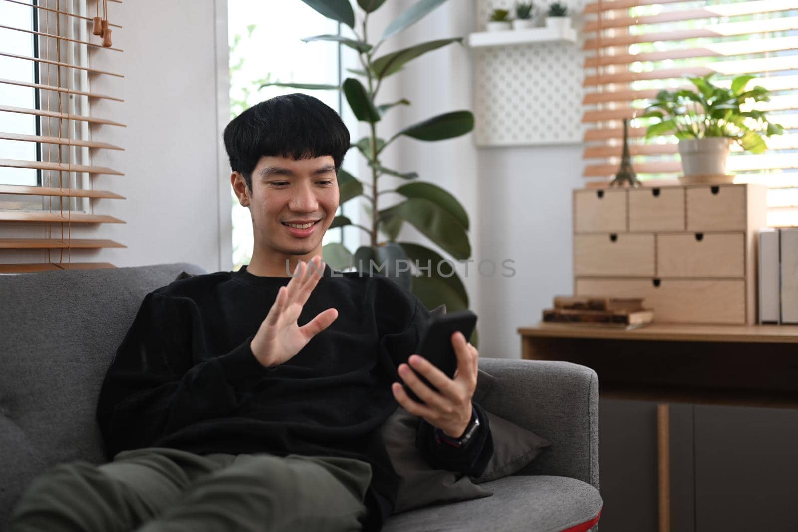 Smiling man sitting on comfortable couch and enjoying chatting online with friends via smart phone.