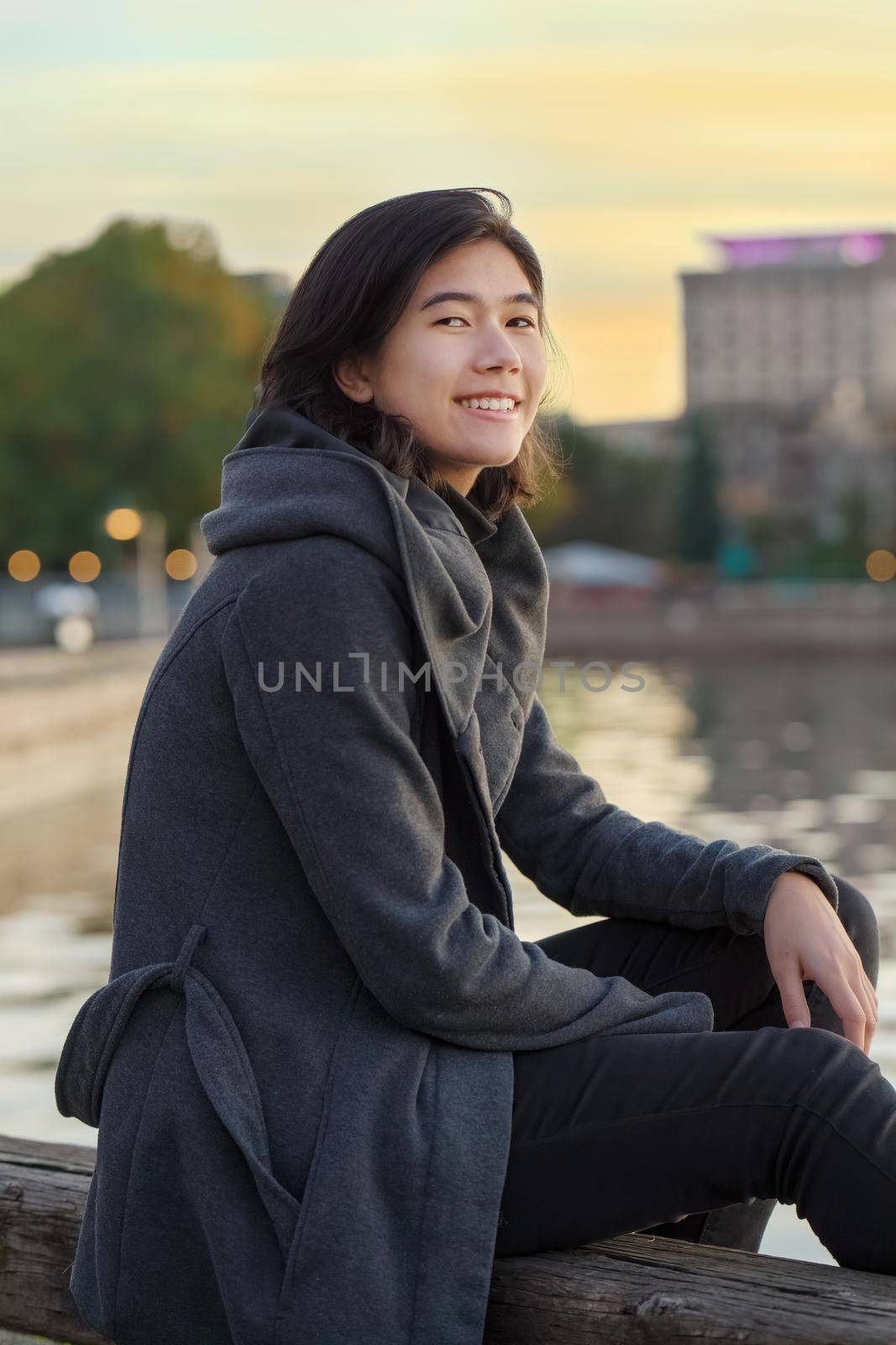 Biracial teen girl sitting by lake at sunset, wearing jacket by jarenwicklund