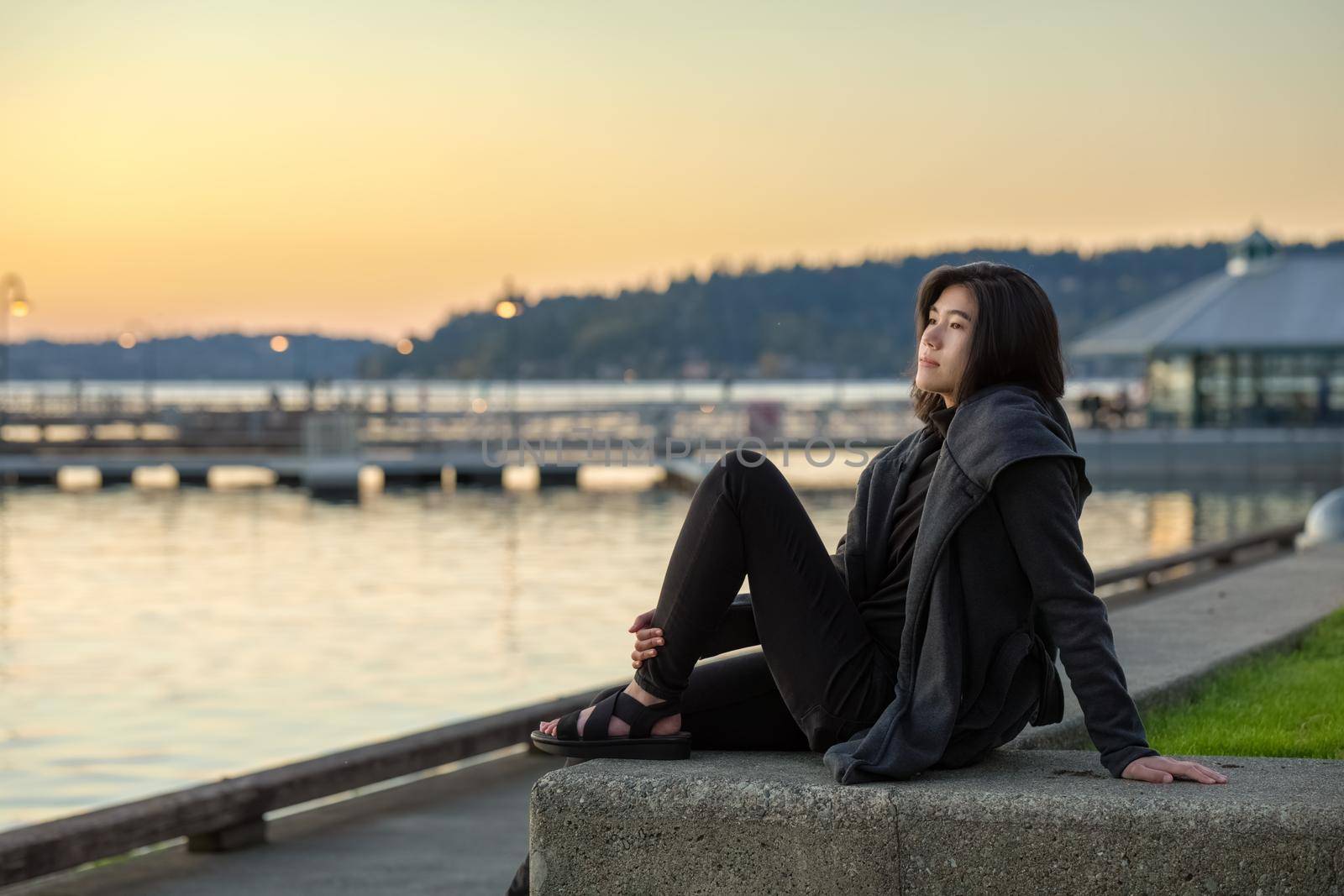 Biracial teen girl sitting by lake at sunset, wearing jacket by jarenwicklund