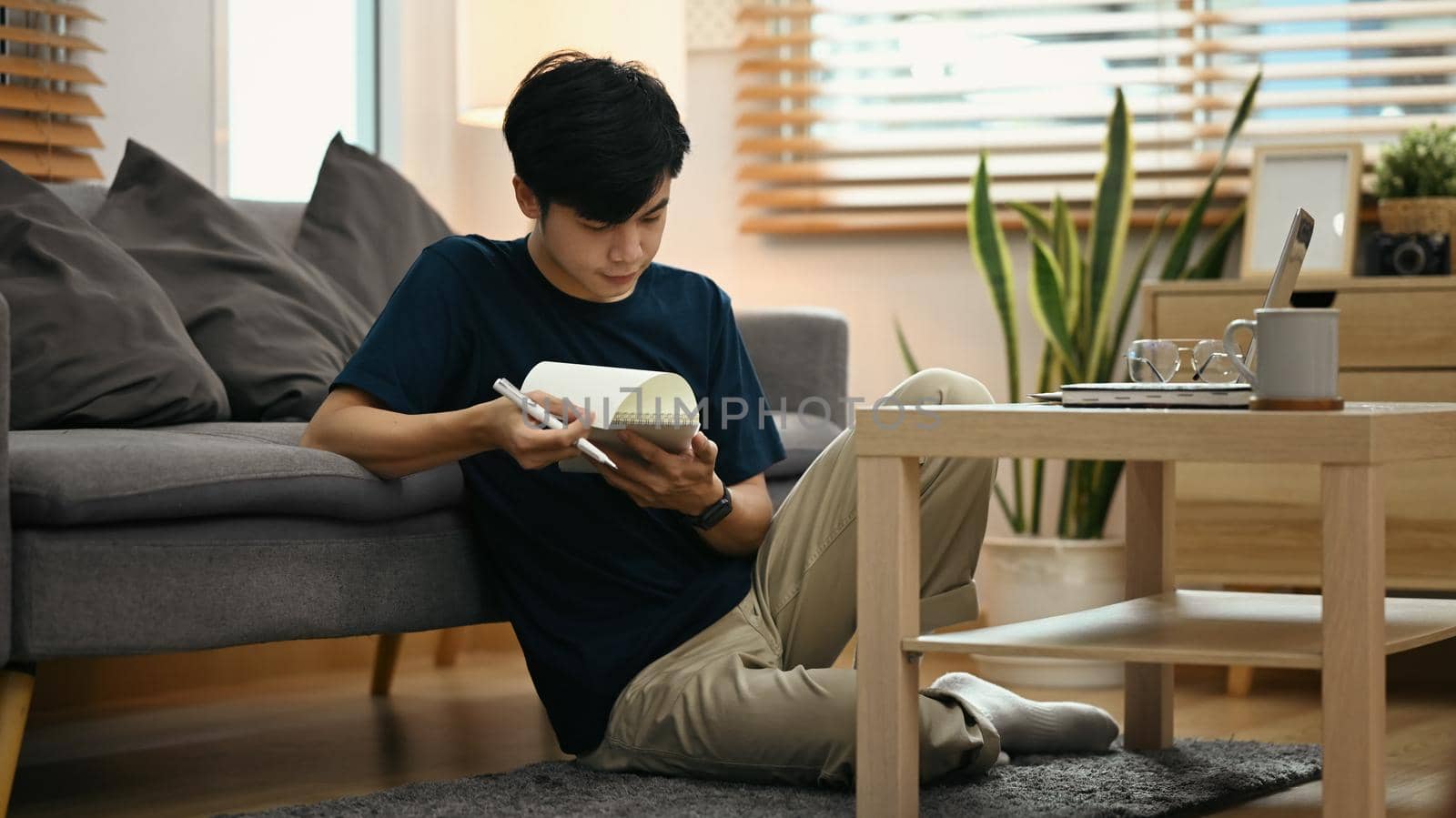 Asian man making notes on notepad and working online from home with laptop while sitting on floor in living room.