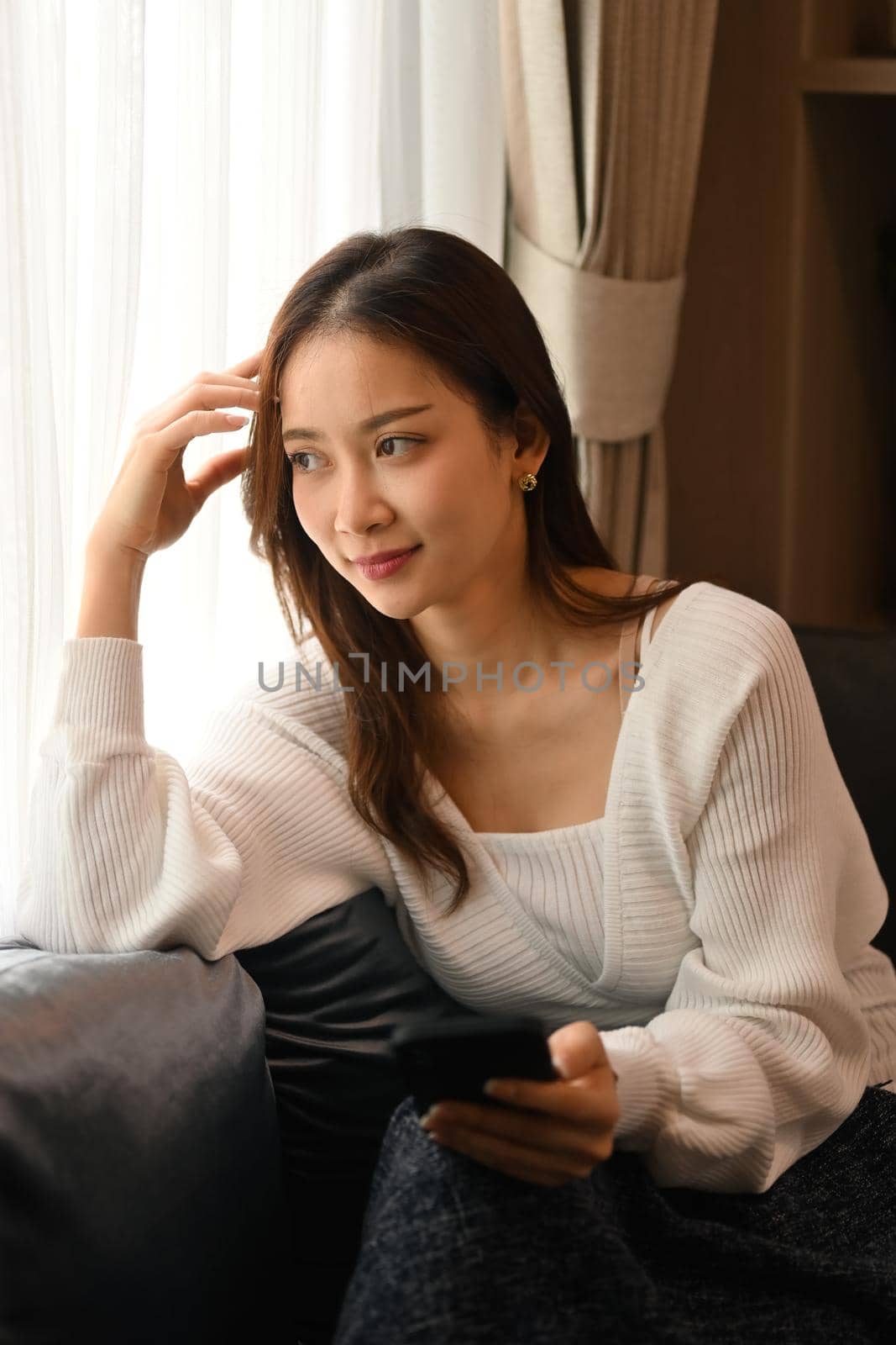 Satisfied young woman resting on couch and using smart phone spending time in winter or autumn weekend.