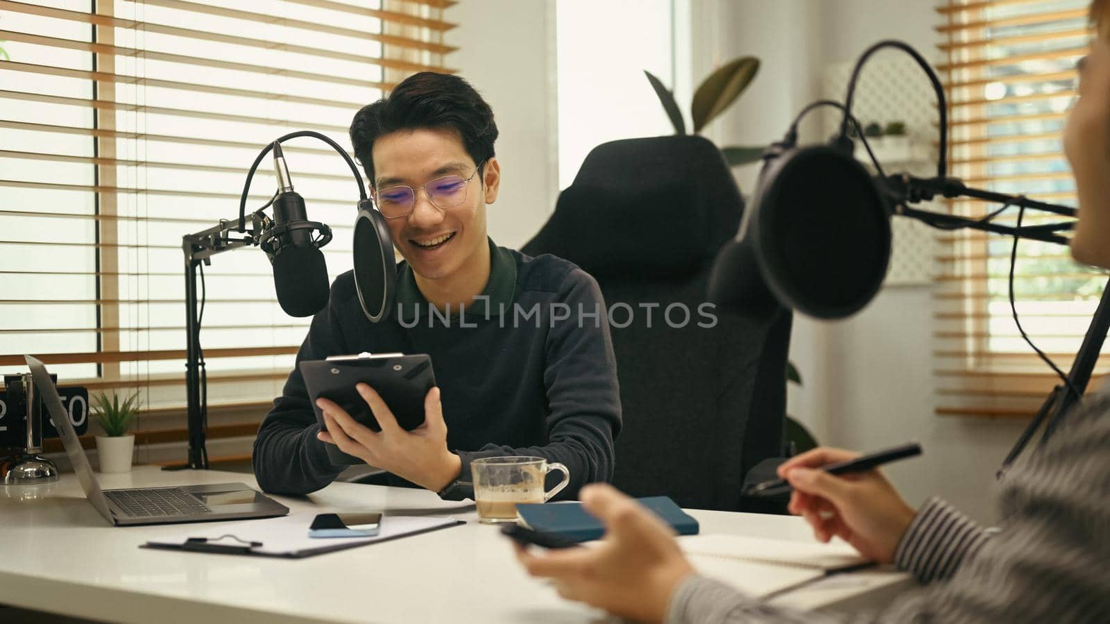 Friendly radio host using condenser microphone for recording voice over radio interview guest conversation at home studio.