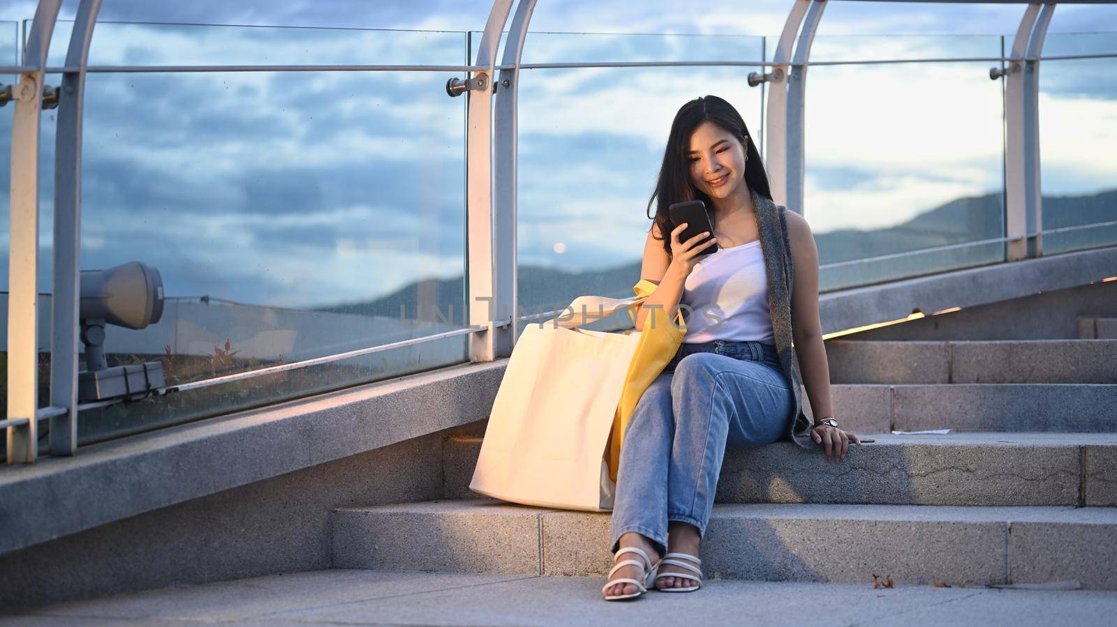 Pretty asian woman sitting using mobile phone and sitting on stairs at terrace with beautiful evening sky in background.