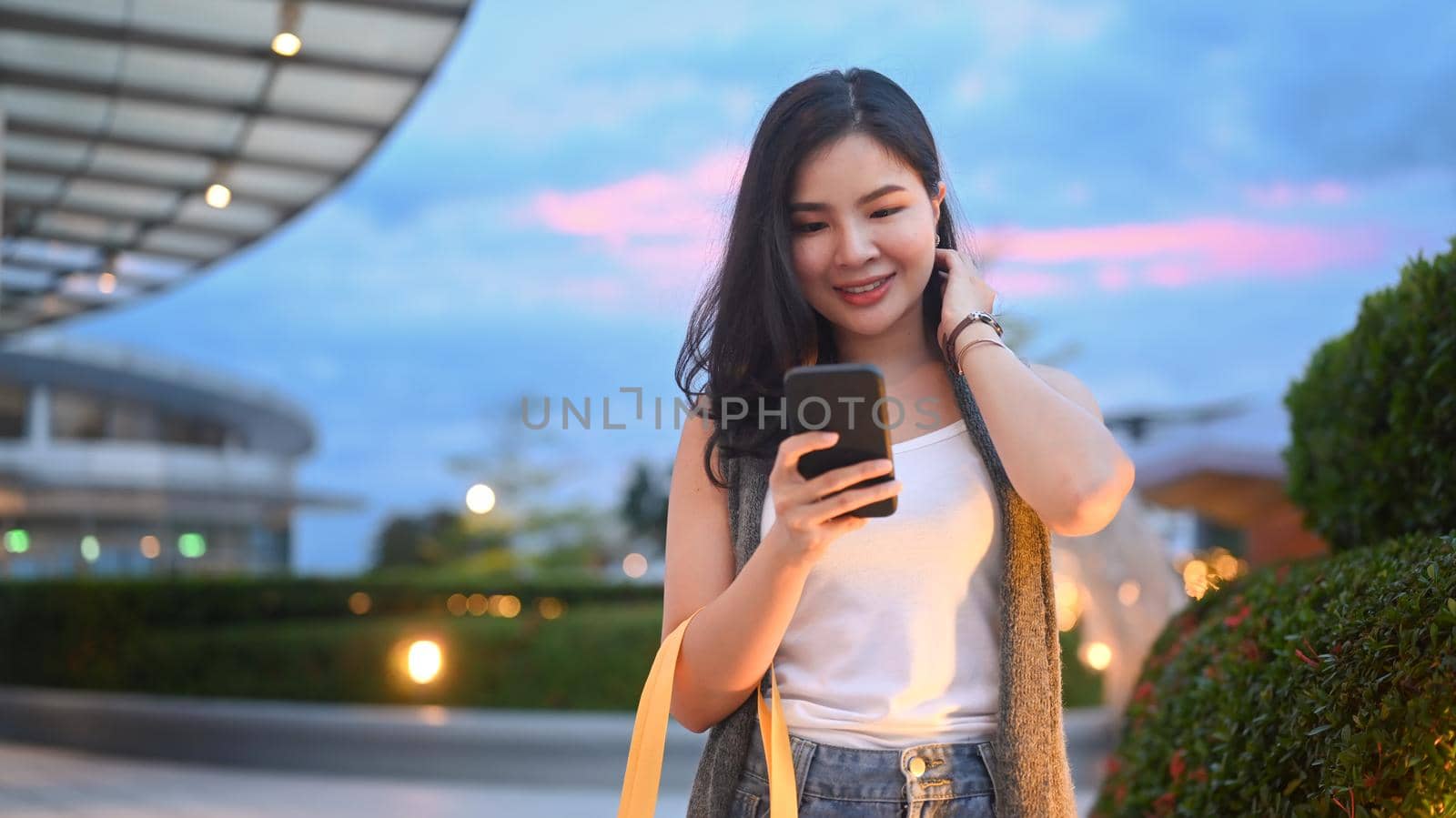 Trendy asian woman using smart phone walking on evening city streets with beautiful evening sky in background.