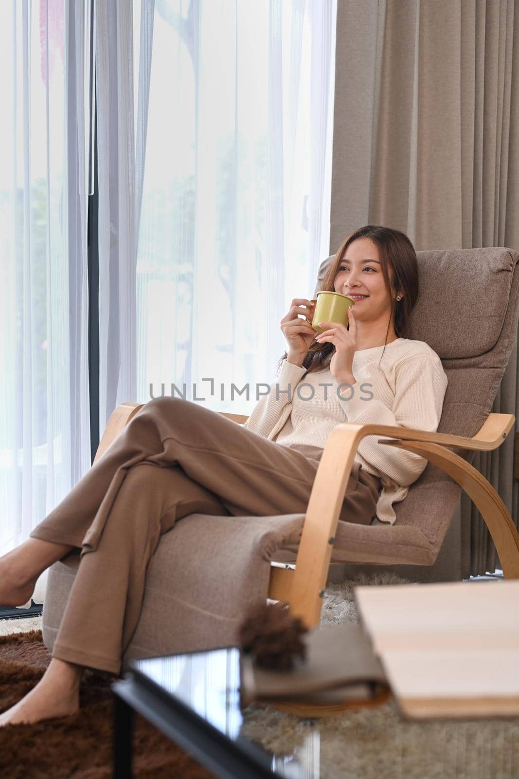 Pleasant woman wearing sweater drinking hot coffee on armchair at home in bright winter morning.