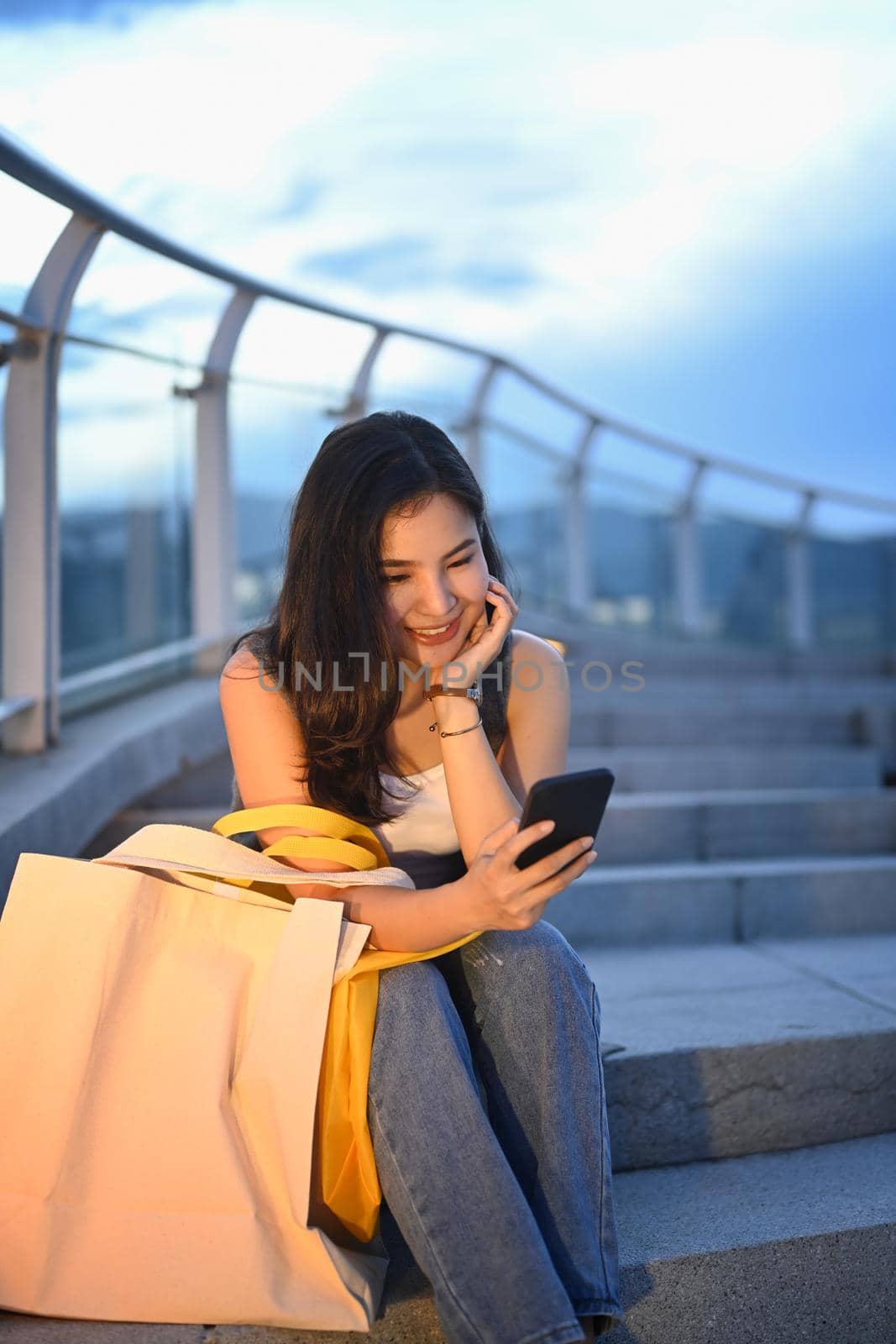 Stylish young woman using mobile phone while sitting on stairs with beautiful evening sky in background.
