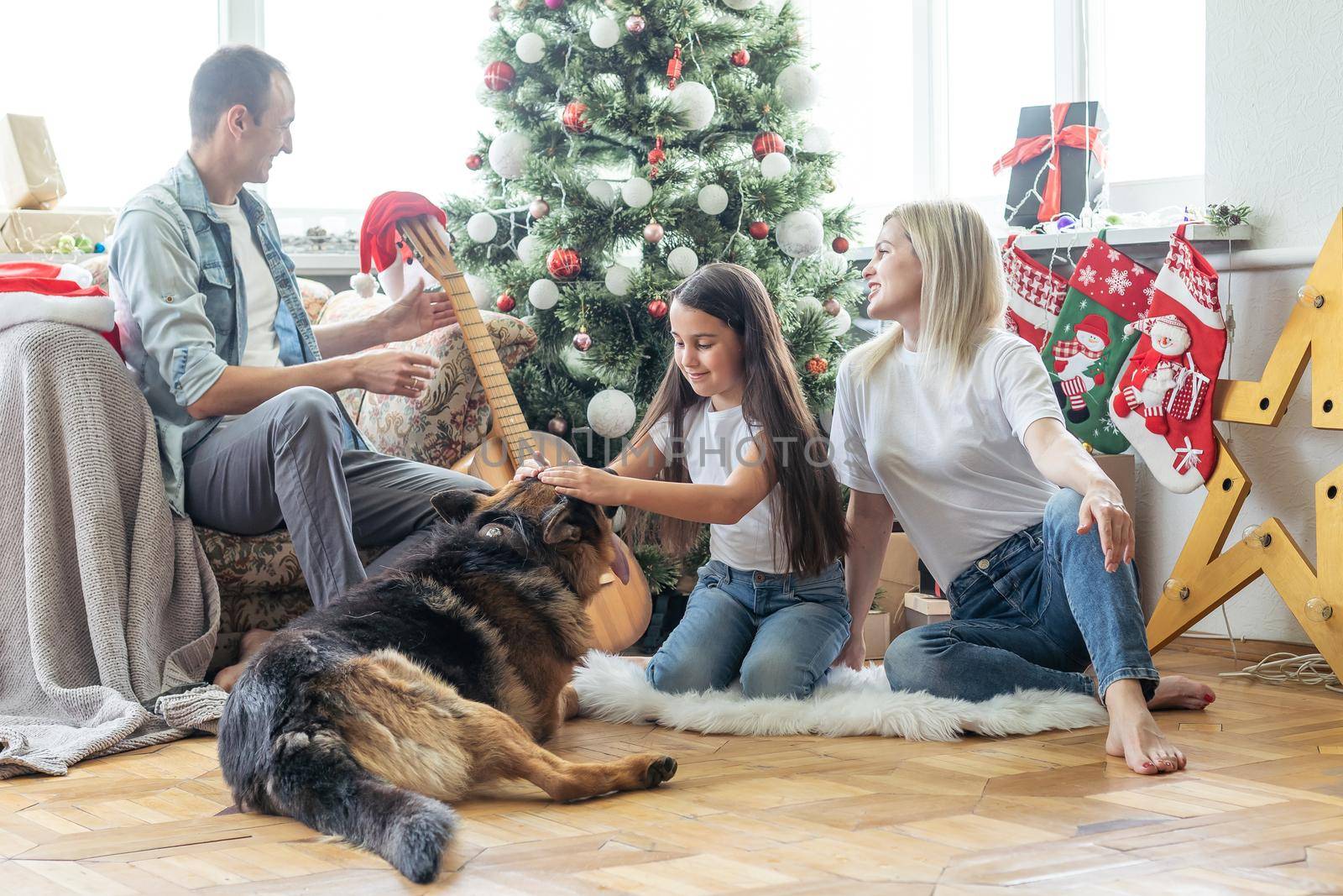 Friendly family is playing with dog near Christmas tree. They are sitting and laughing.