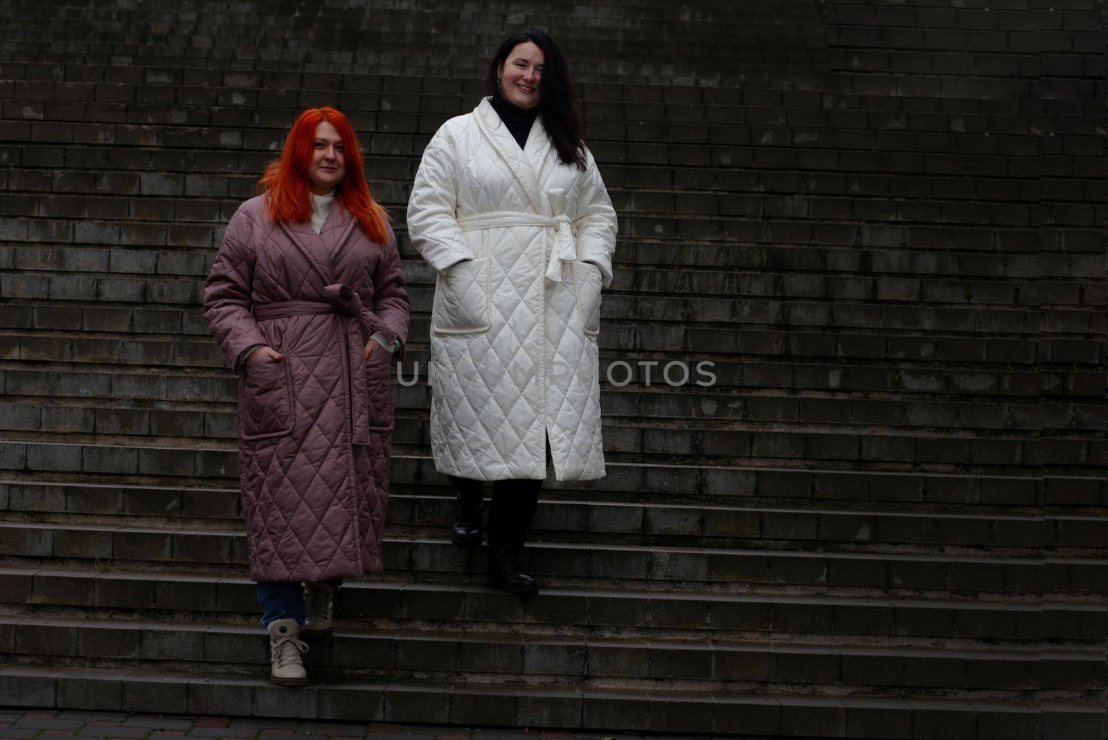 Woman in white coat and girl in brown coat standing on a stone tem. Two plus size women posing in winter clothes outdoors.
