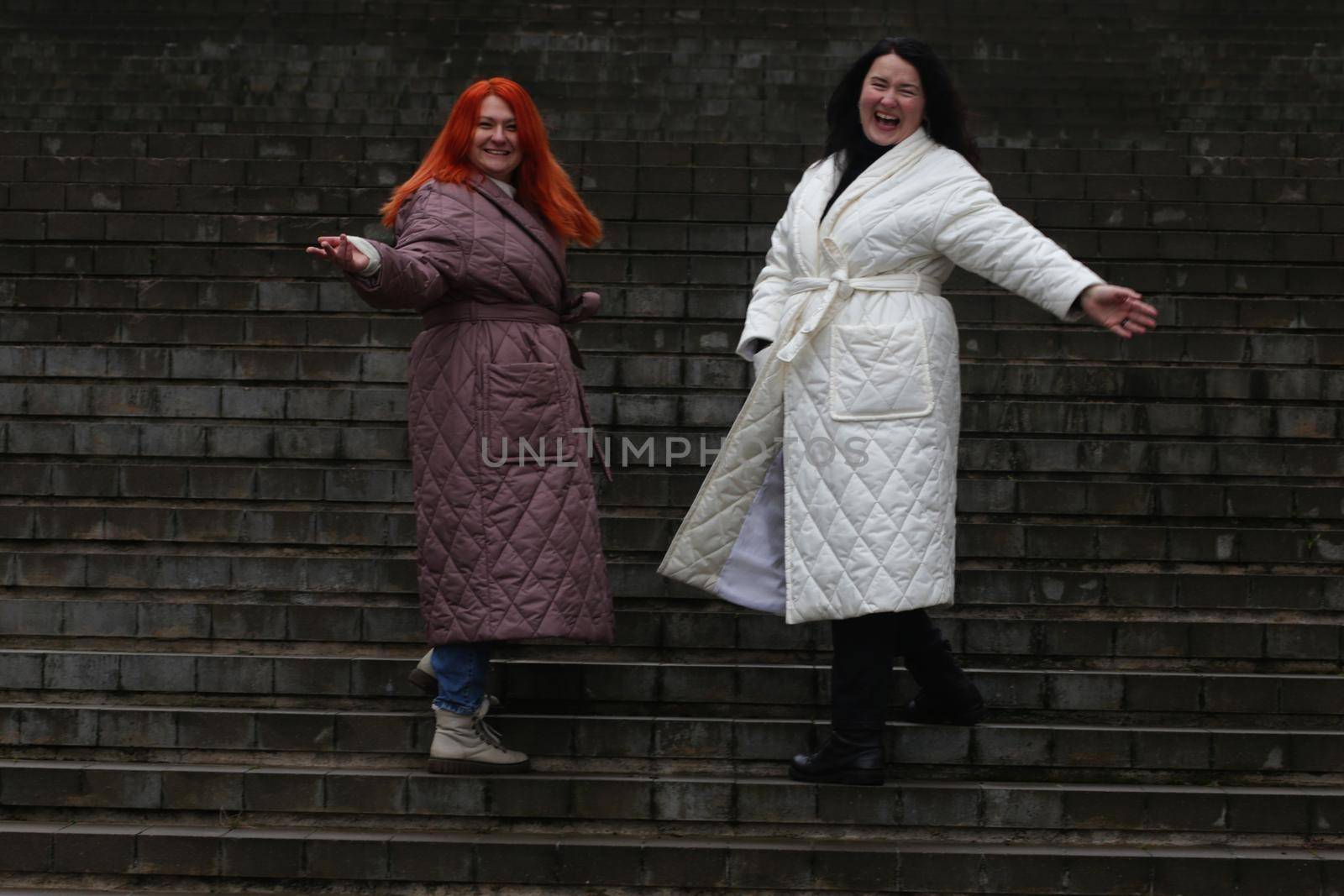 Two emotional plus size women in a white and red coat, a brown-haired woman and a brunette, against the background of an old dark gray concrete staircase.