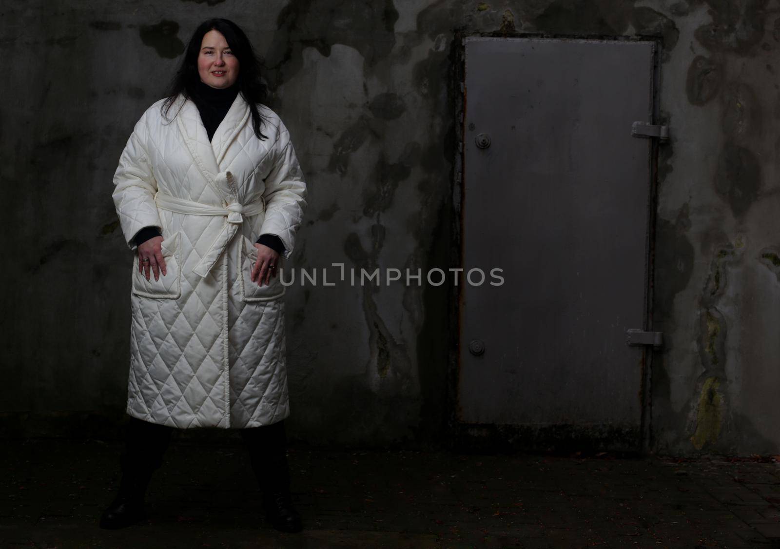 Portrait of a woman plus size model with dark hair in a white XXL winter coat and black trousers posing against the wall.