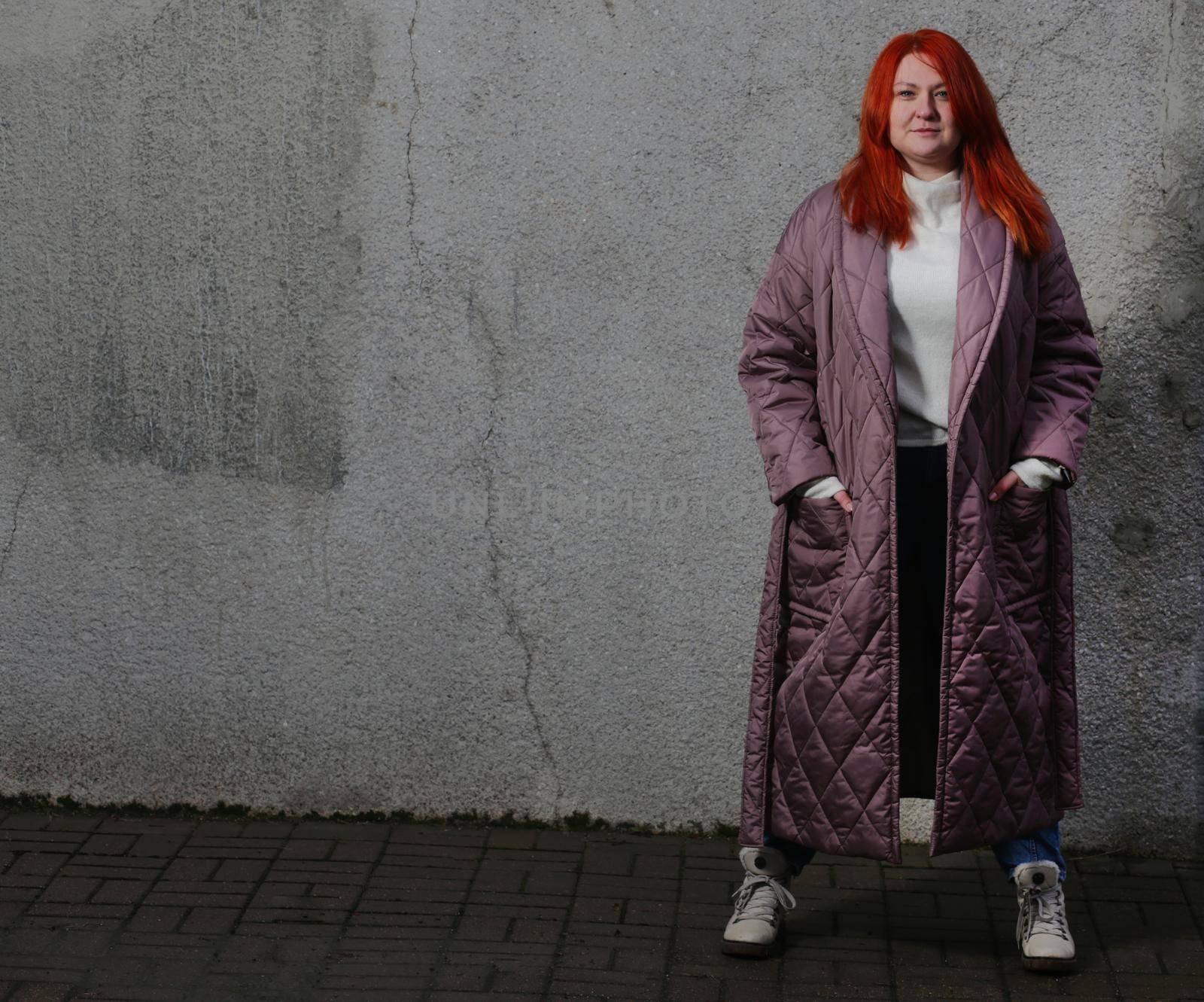 Image of a girl with red hair in a long brown coat holding her hands in her pockets against the background of an old ramshackle concrete wall. Girl in white sweater and white boots in a brown long coat looking at the camera.