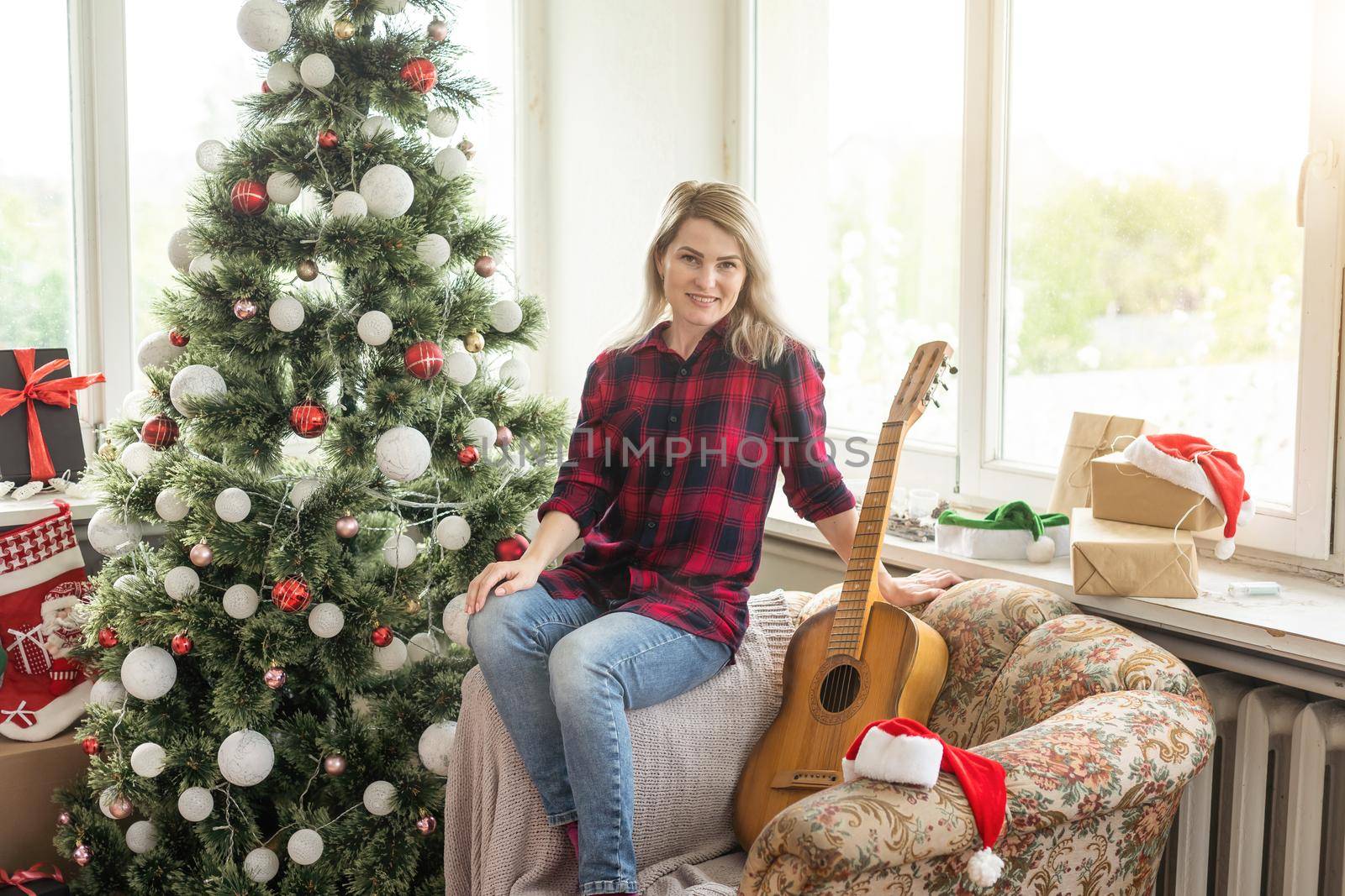 beautiful girl sits near the Christmas tree decorated with balls and light and playing the guitar and singing. Young beautiful smiling woman play guitar. by Andelov13