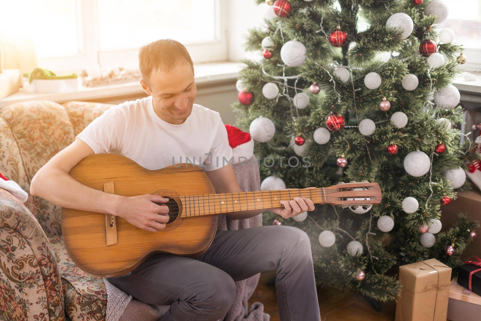 young handsome man sitting on the sofa, he is playing guitar, music, christmas, relax concept by Andelov13