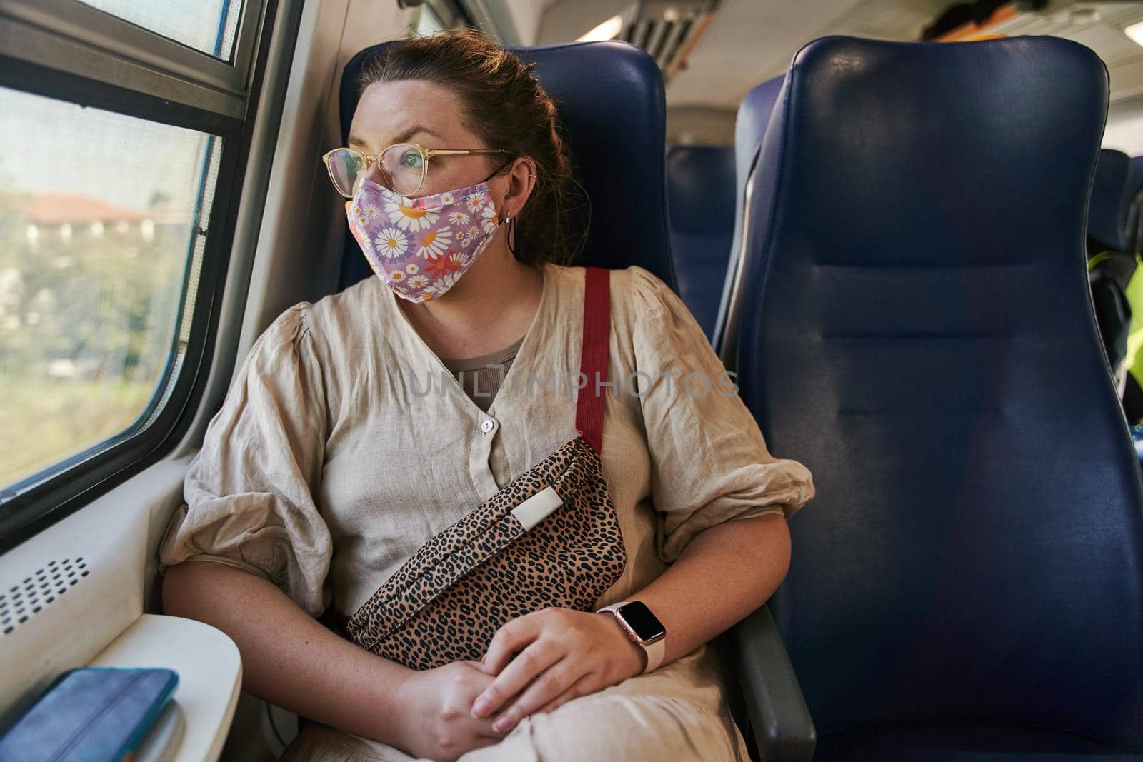 A girl in glasses and a medical mask riding a train and looking out the window. High quality photo