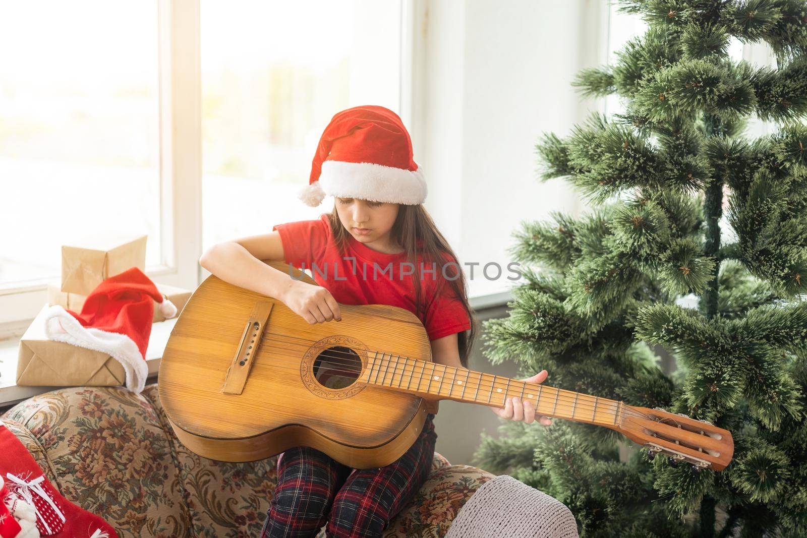 Happy little girl in red hat playing the on the background of christmas tree. Talent kid having fun on winter holidays at cozy decorated home. by Andelov13