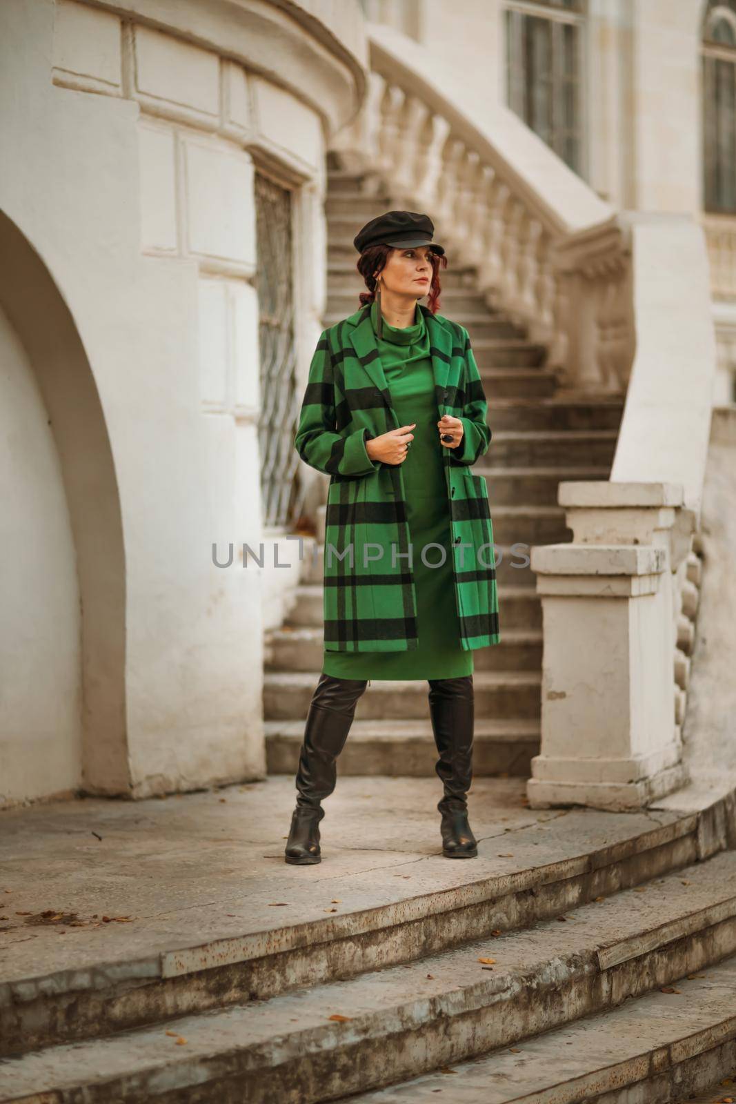 Outdoor fashion portrait of an elegant fashionable brunette woman, model in a stylish cap, green dress, posing at sunset in a European city in autumn