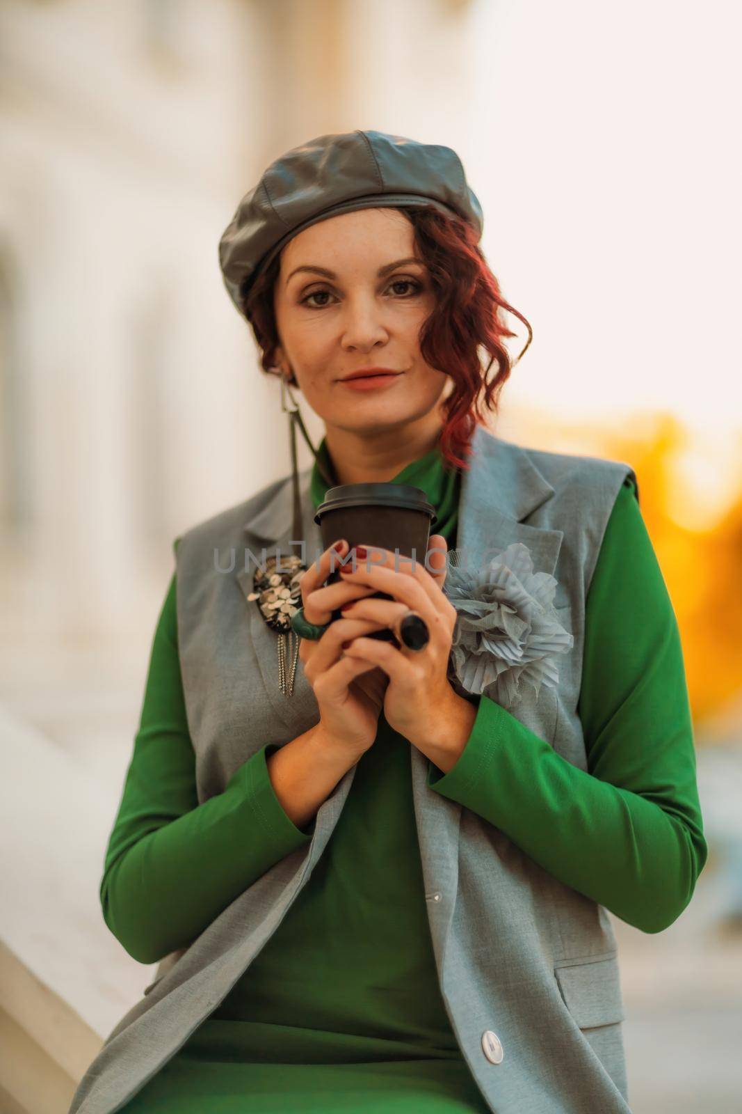 Outdoor fashion portrait of an elegant fashionable brunette model in a beret, green dress and a gray waistcoat posing at sunset in a european city in autumn. by Matiunina