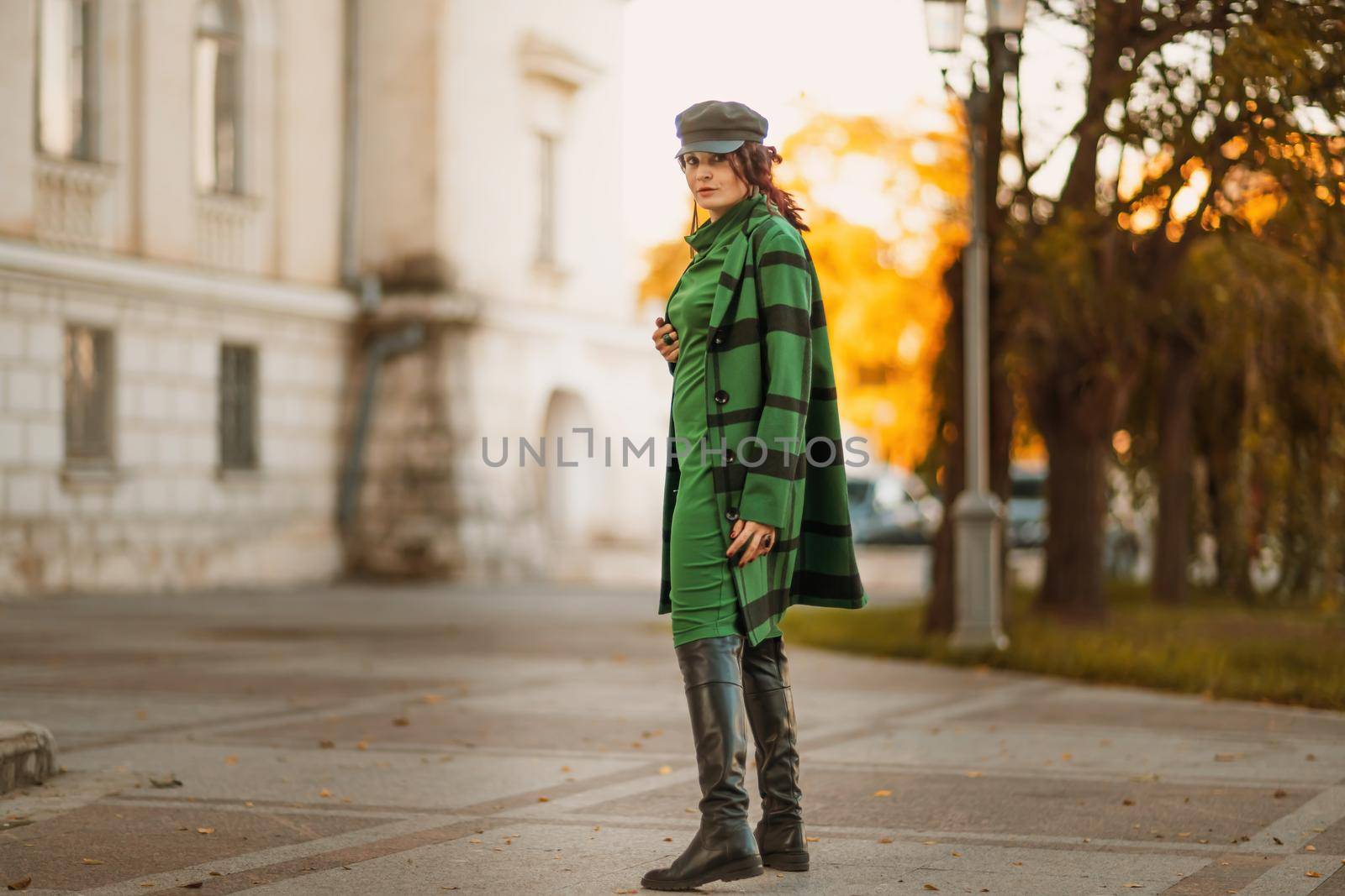 Outdoor fashion portrait of an elegant fashionable brunette woman, model in a stylish cap, green dress, posing at sunset in a European city in autumn