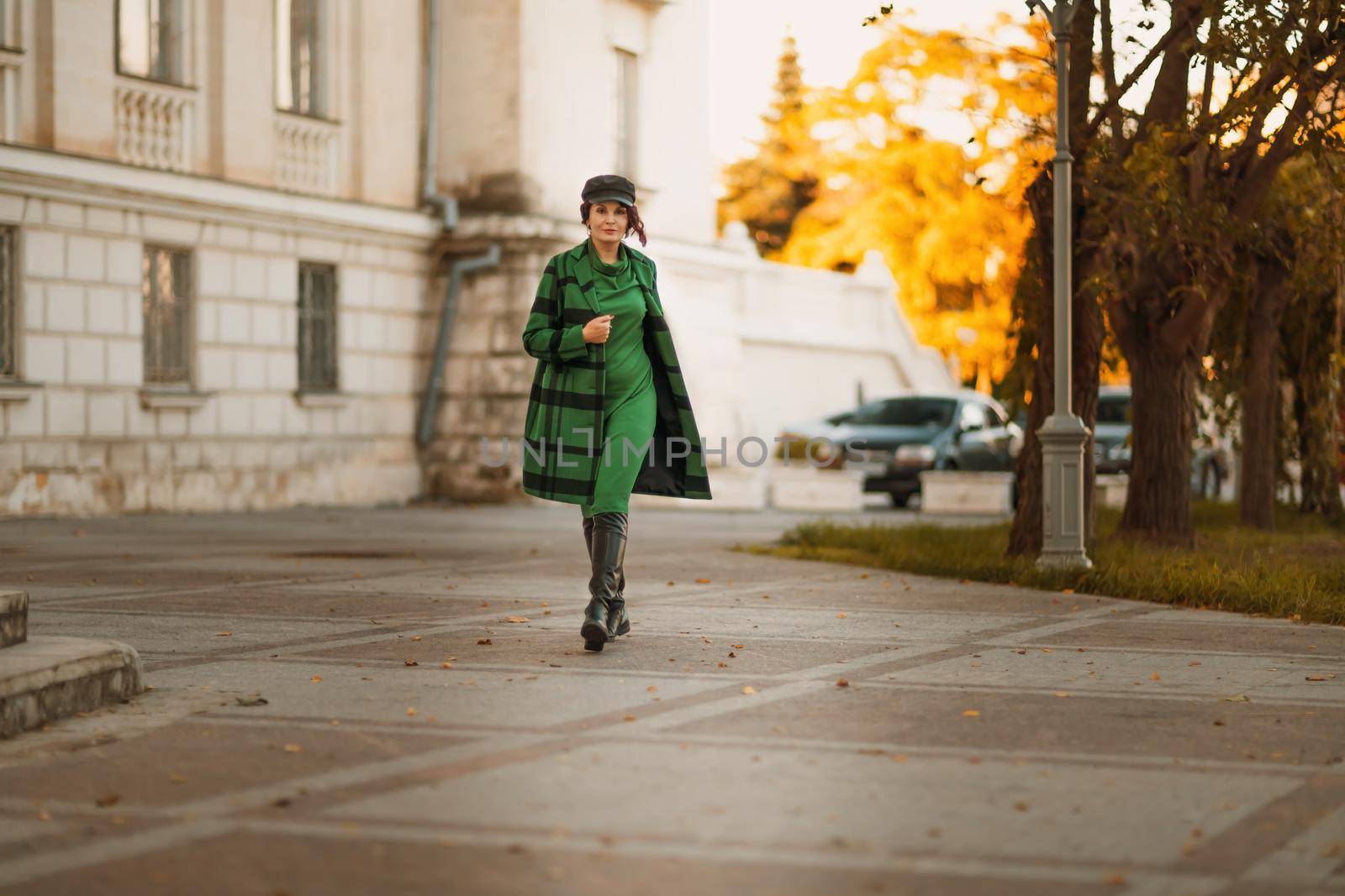 Outdoor fashion portrait of an elegant fashionable brunette woman, model in a stylish cap, green dress, posing at sunset in a European city in autumn