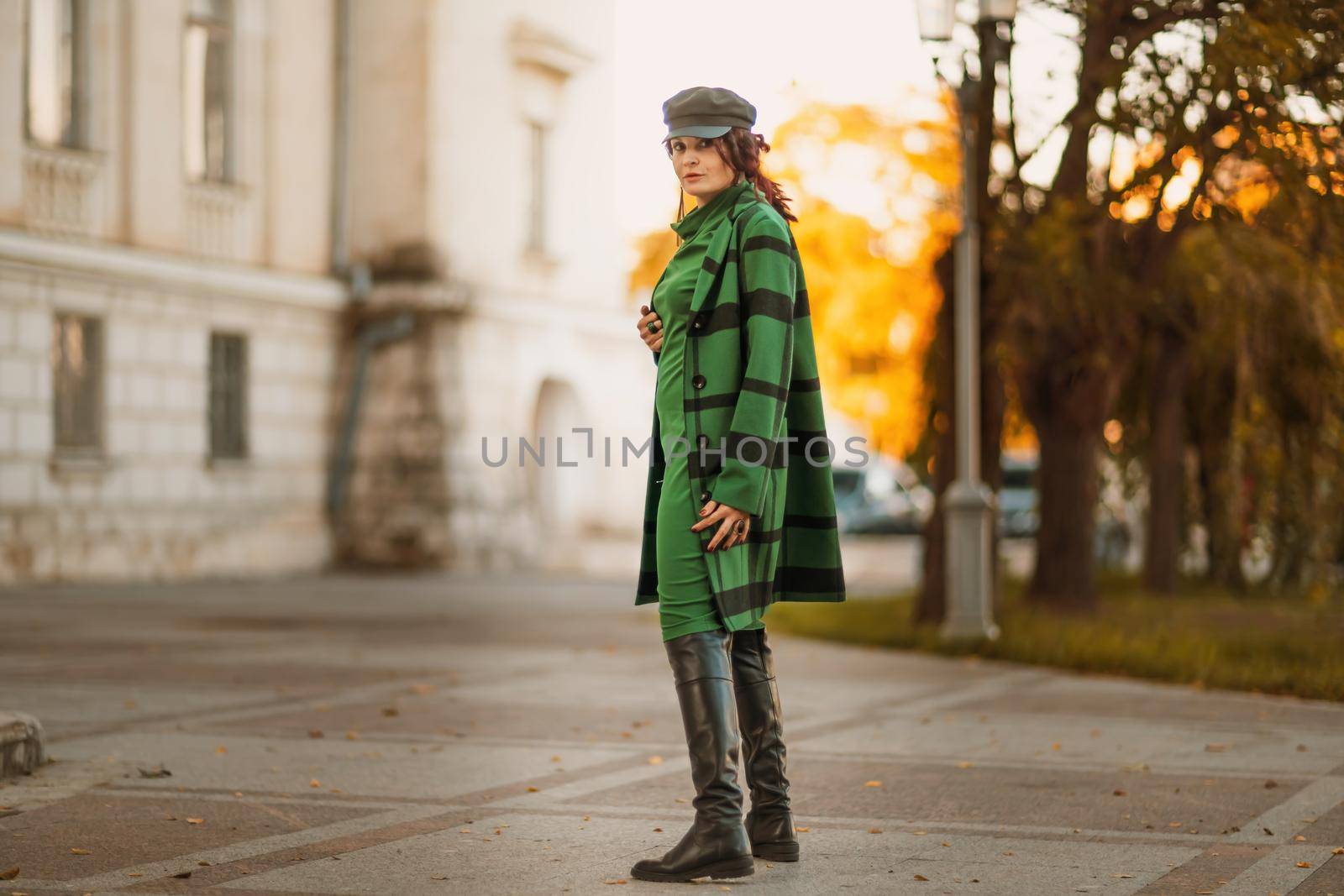 Outdoor fashion portrait of an elegant fashionable brunette woman, model in a stylish cap, green dress, posing at sunset in a European city in autumn