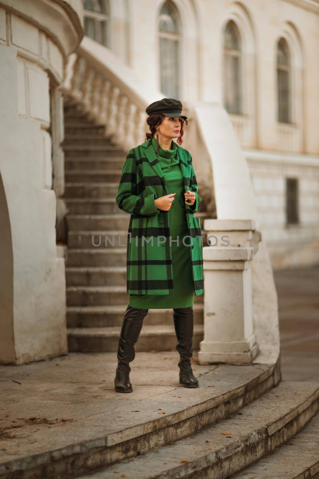 Outdoor fashion portrait of an elegant fashionable brunette woman, model in a stylish cap, green dress, posing at sunset in a European city in autumn