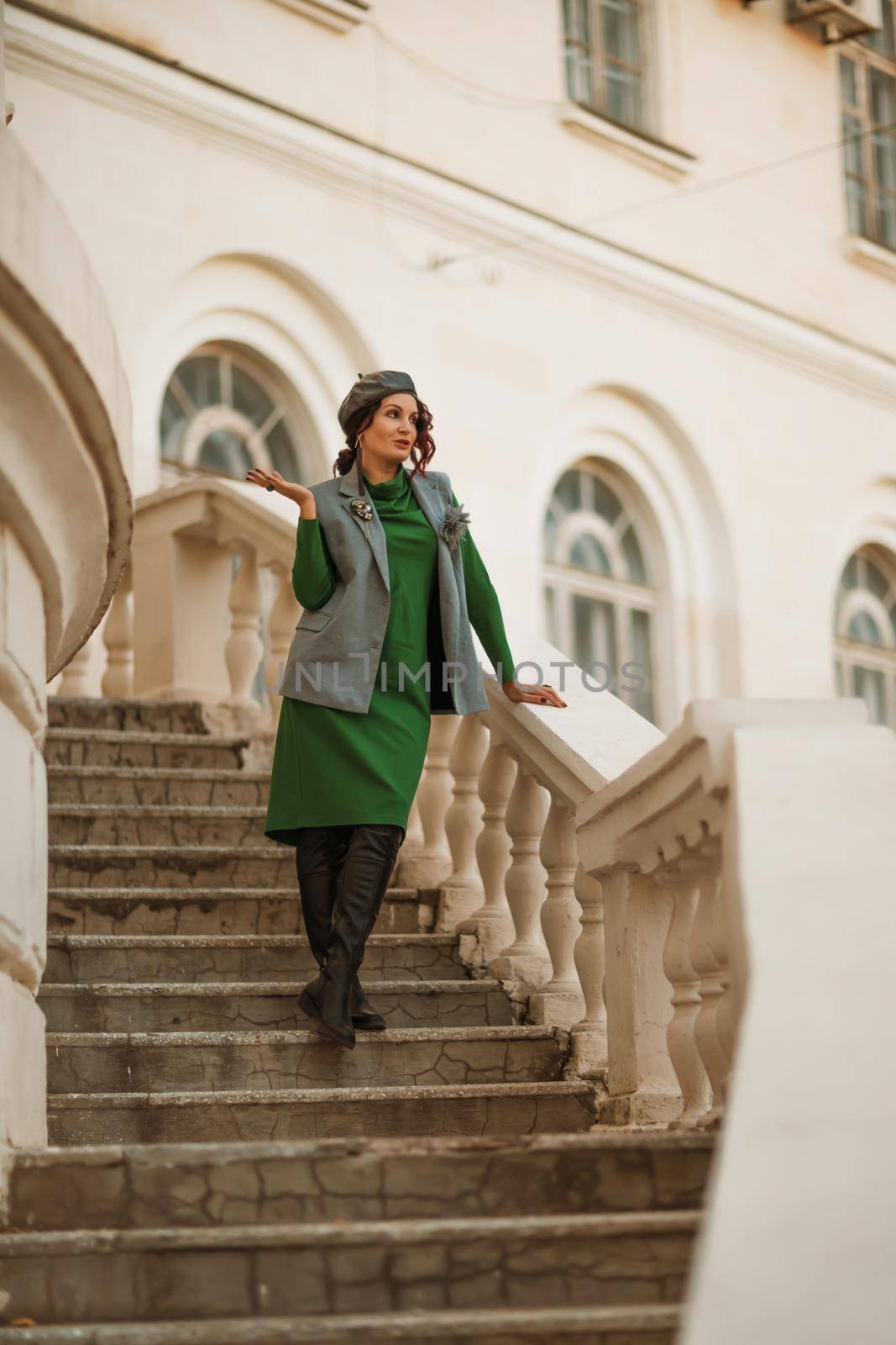 Outdoor fashion portrait of an elegant fashionable brunette model in a beret, green dress and a gray waistcoat posing at sunset in a european city in autumn. by Matiunina