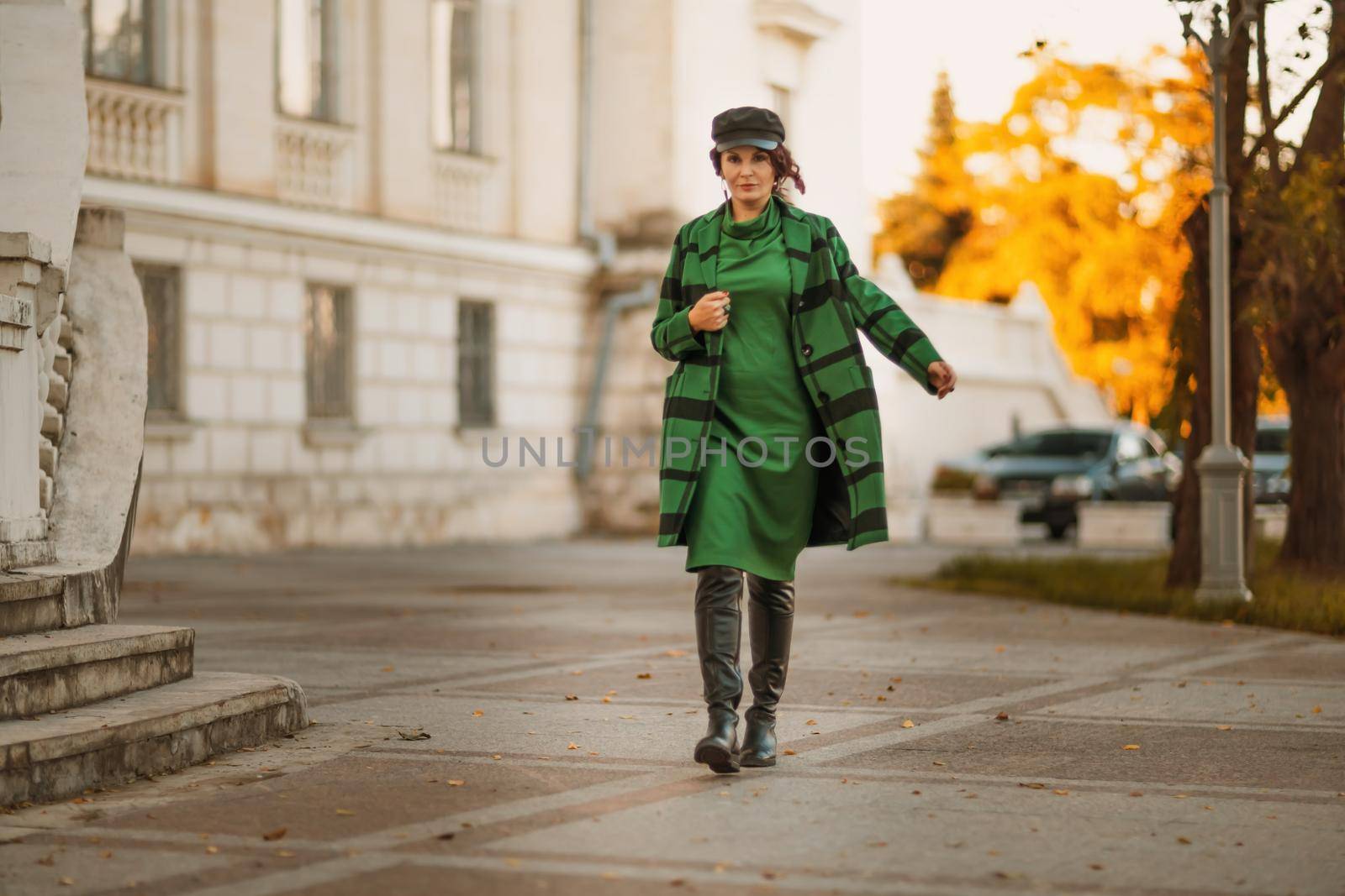 Outdoor fashion portrait of an elegant fashionable brunette woman, model in a stylish cap, green dress, posing at sunset in a European city in autumn