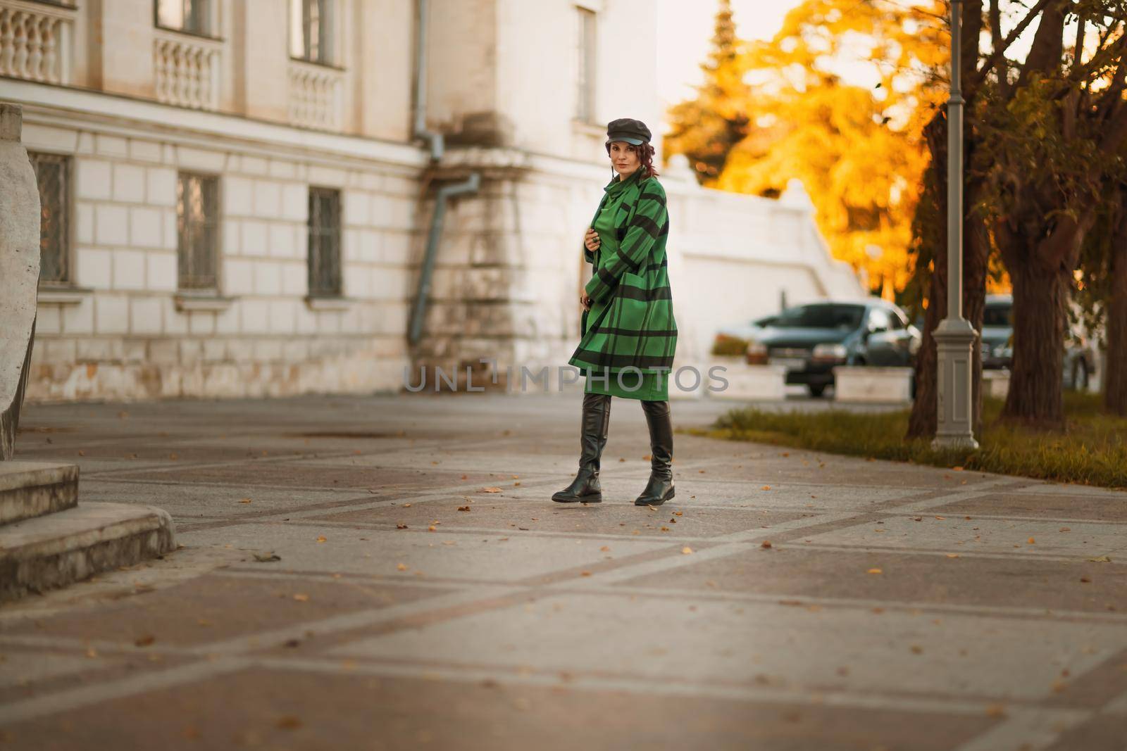 Outdoor fashion portrait of an elegant fashionable brunette woman, model in a stylish cap, green dress, posing at sunset in a European city in autumn