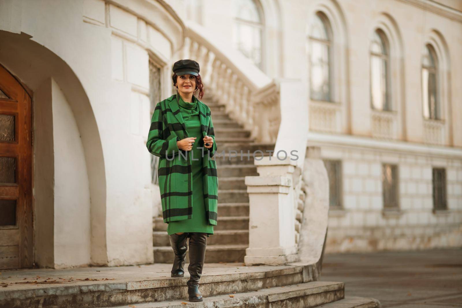 Outdoor fashion portrait of an elegant fashionable brunette woman, model in a stylish cap, green dress, posing at sunset in a European city in autumn