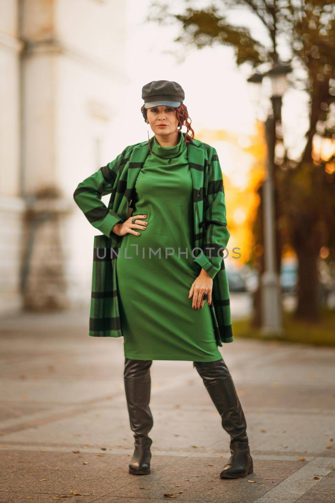 Outdoor fashion portrait of an elegant fashionable brunette woman, model in a stylish cap, green dress, posing at sunset in a European city in autumn