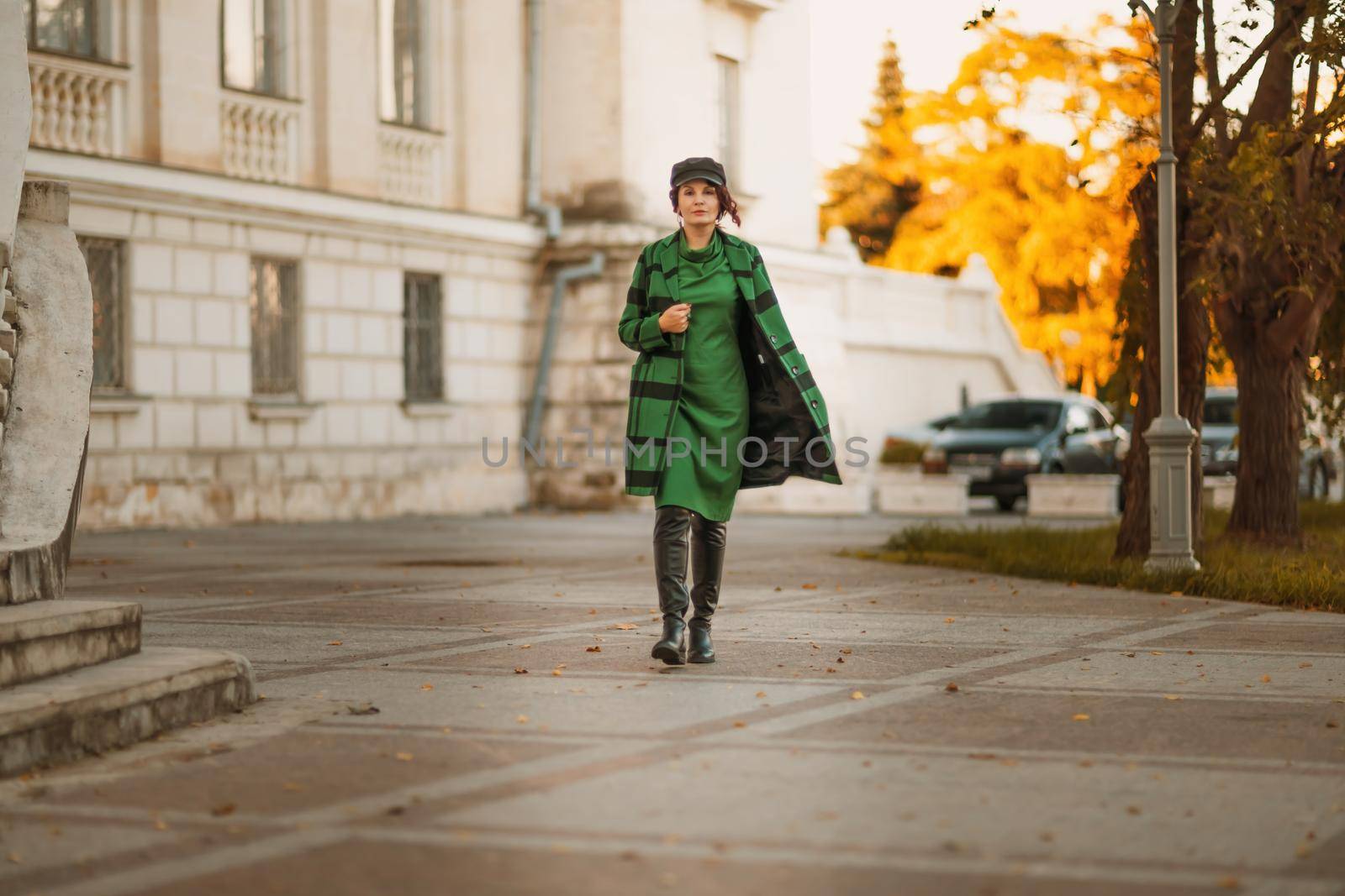 Outdoor fashion portrait of an elegant fashionable brunette woman, model in a stylish cap, green dress, posing at sunset in a European city in autumn