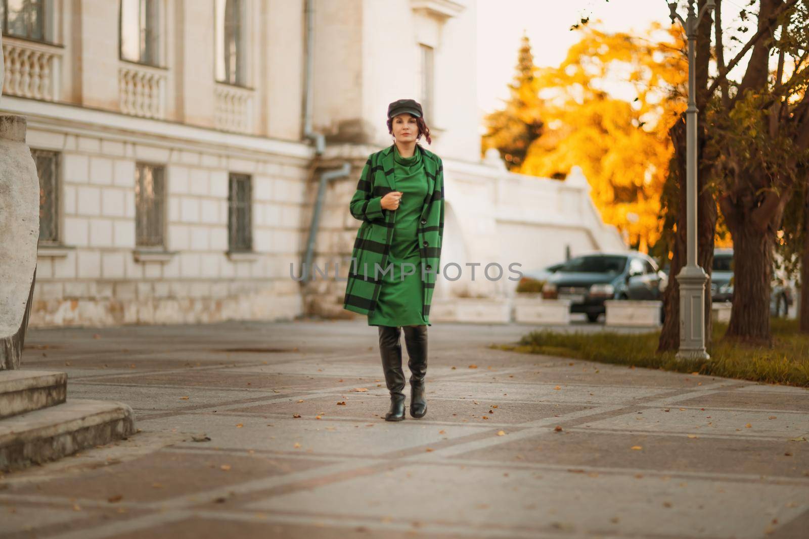 Outdoor fashion portrait of an elegant fashionable brunette woman, model in a stylish cap, green dress, posing at sunset in a European city in autumn