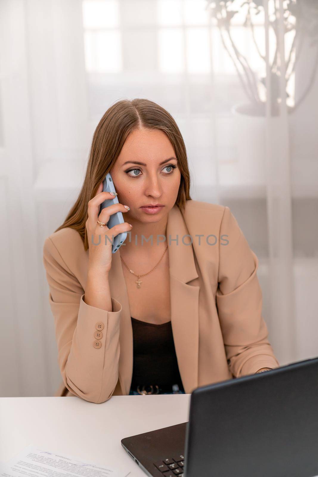 European professional woman is sitting with a laptop at a table in a home office, a positive woman is studying while working on a PC. She is wearing a beige jacket and jeans and is on the phone