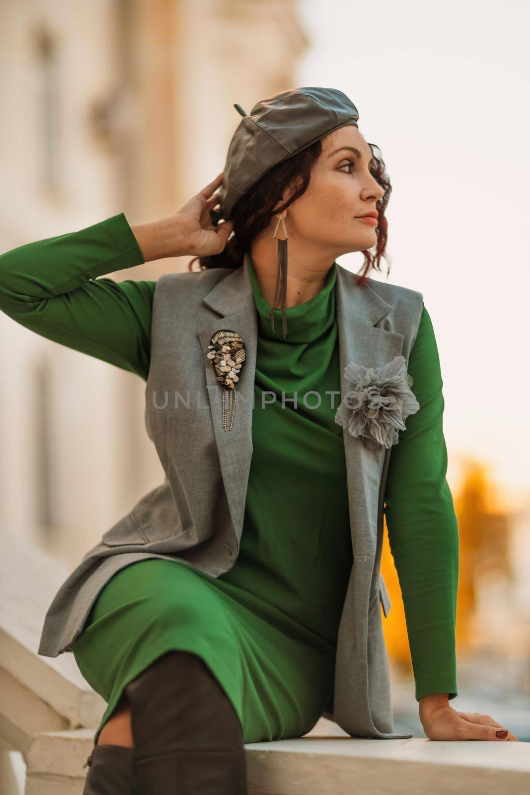 Outdoor fashion portrait of an elegant fashionable brunette model in a beret, green dress and a gray waistcoat posing at sunset in a european city in autumn