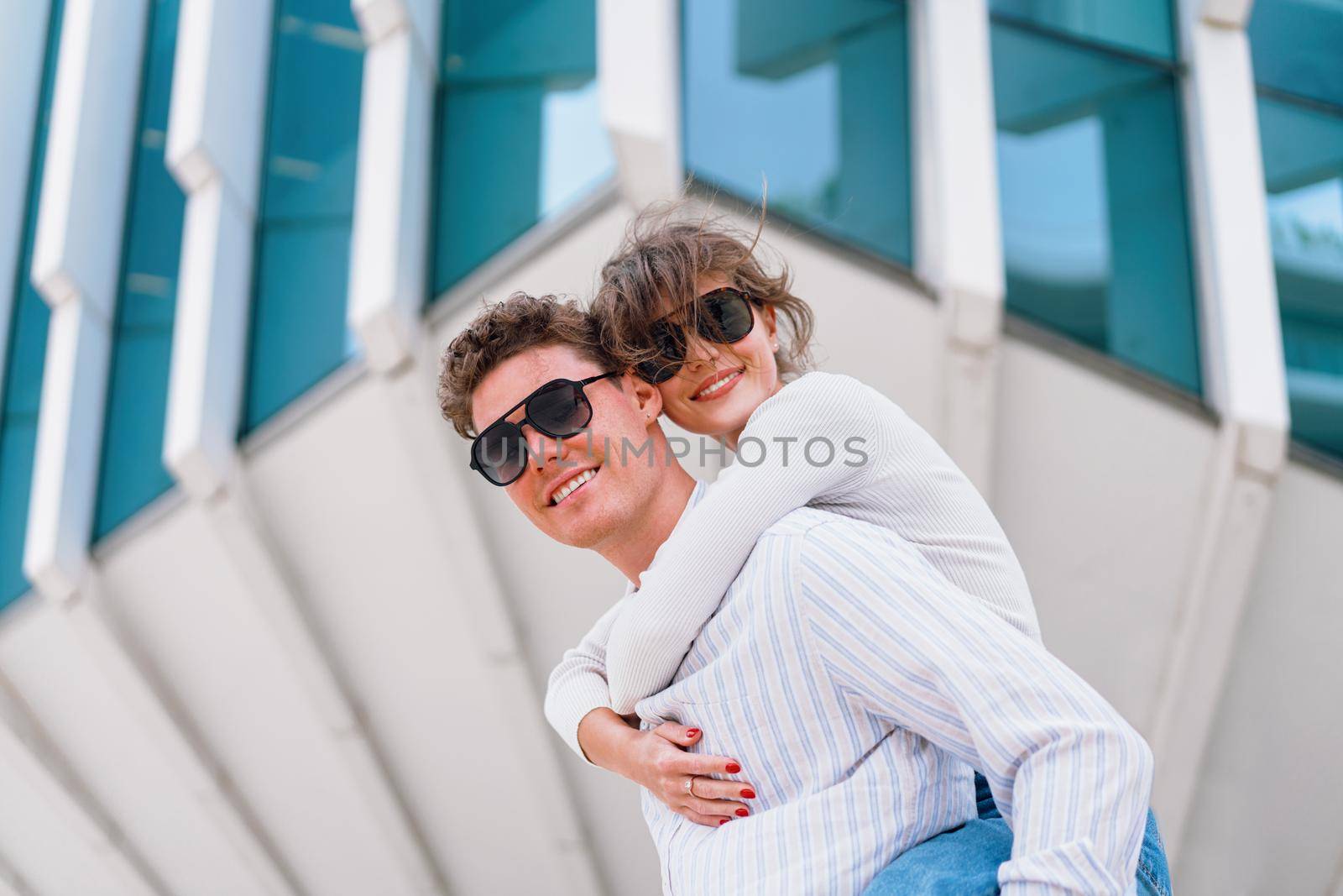 Carefree young urban couple in sunglasses doing piggyback at city street outdoor with modern commercial building on background. Man carrying young woman on his back, positive emotion