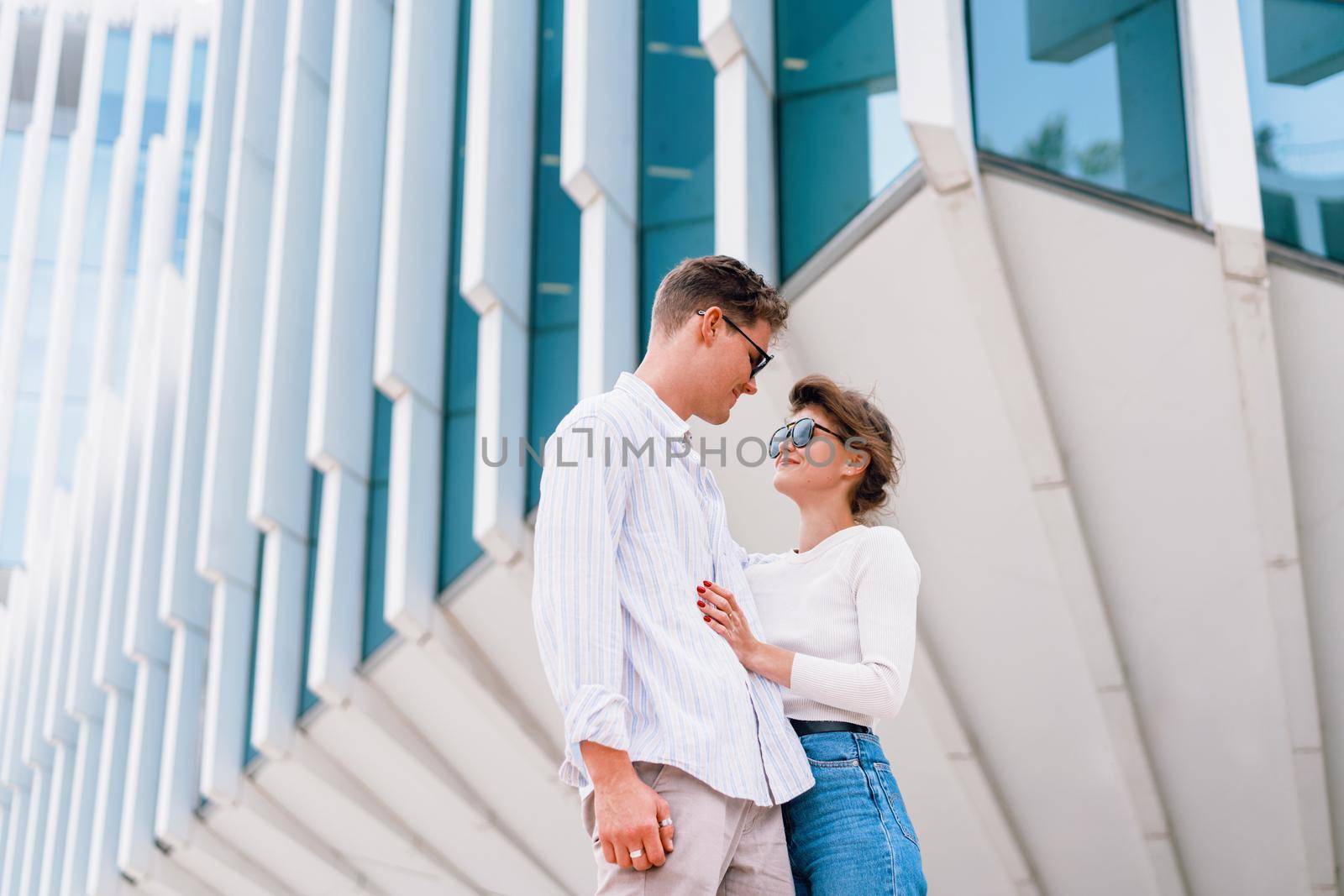 Young beautiful hipster couple in sunglasses in love walking on modern city street by andreonegin