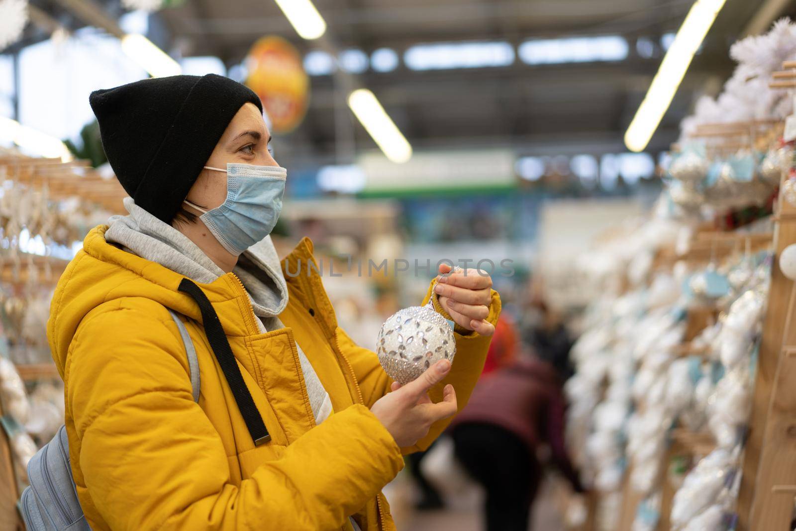 Christmas decoration shopping. Caucasian woman choose new year decor in shopping mall. Medium age female dressed medical mask and yellow winter parka buying Christmas ornament toys indoor store