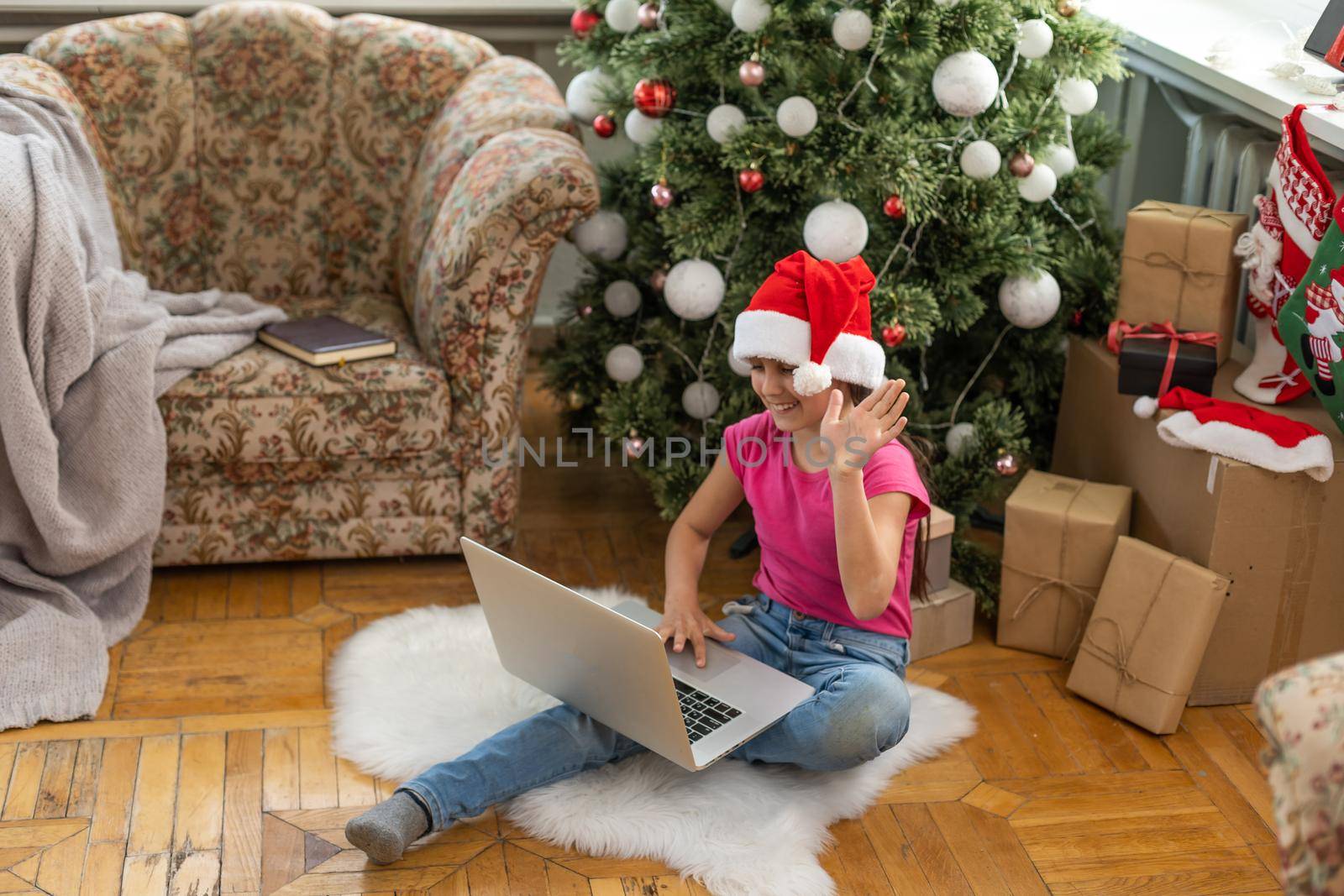A happy little girl opens a gift under the Christmas tree at home and communicates with her grandparents via a laptop via video link. Holiday concept for Christmas and New Year at home by Andelov13