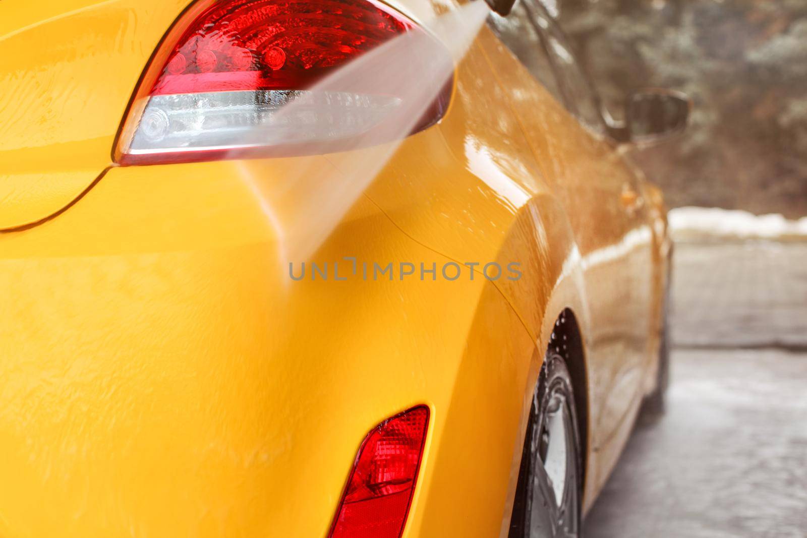 Yellow car rear light and bumper washed in self serve carwash, water jet spraying on gloss surface.