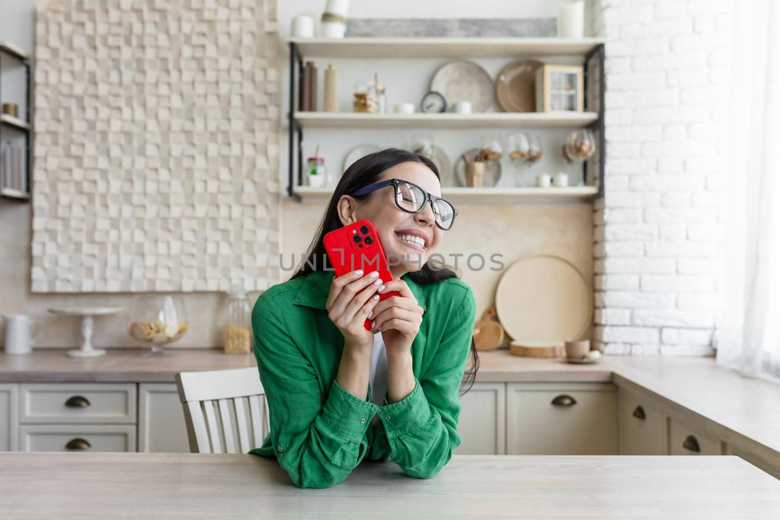 In love and happy young beautiful woman sends and receives love messages on the red phone from a loved one, from a boyfriend. Standing in the kitchen at home in glasses and a green shirt.